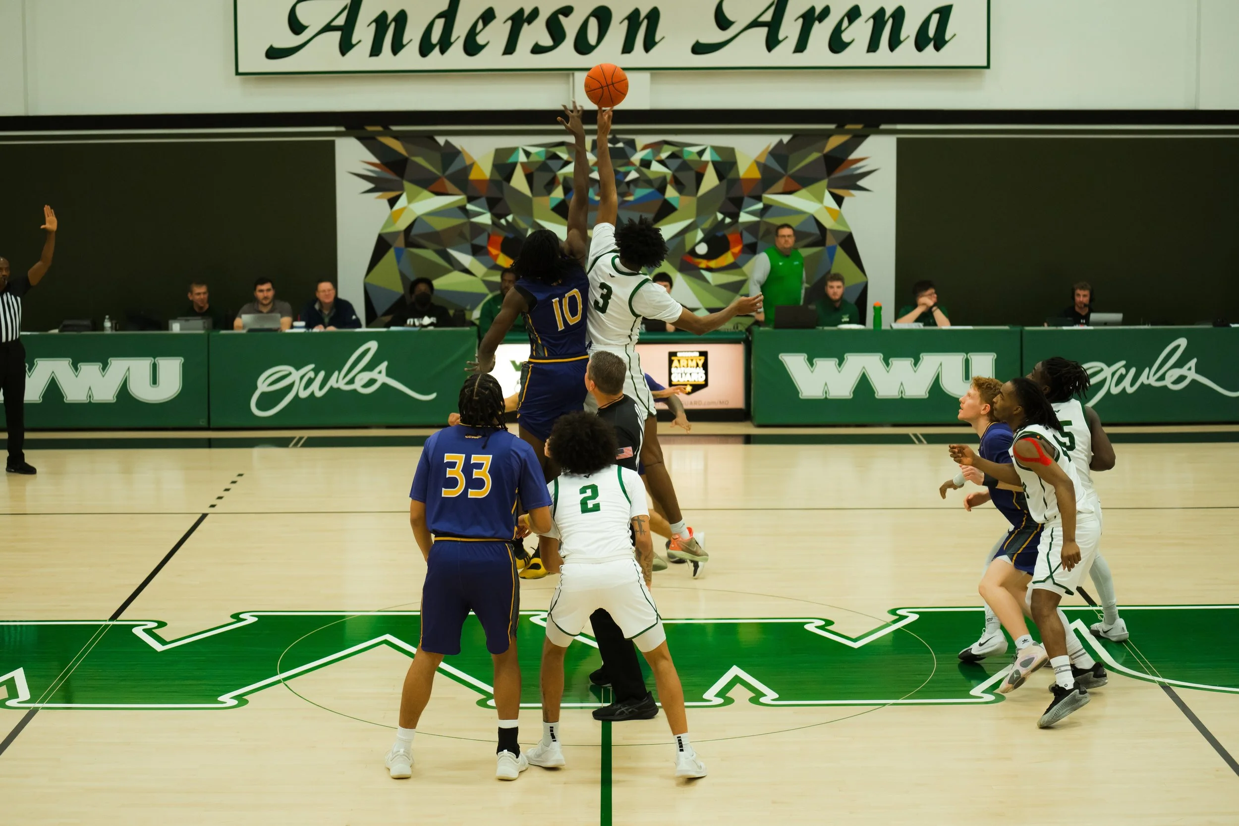A basketball game starting at Anderson Arena with players jumping for the opening tip-off, surrounded by officials and spectators, with a colorful tiger mural in the background.
