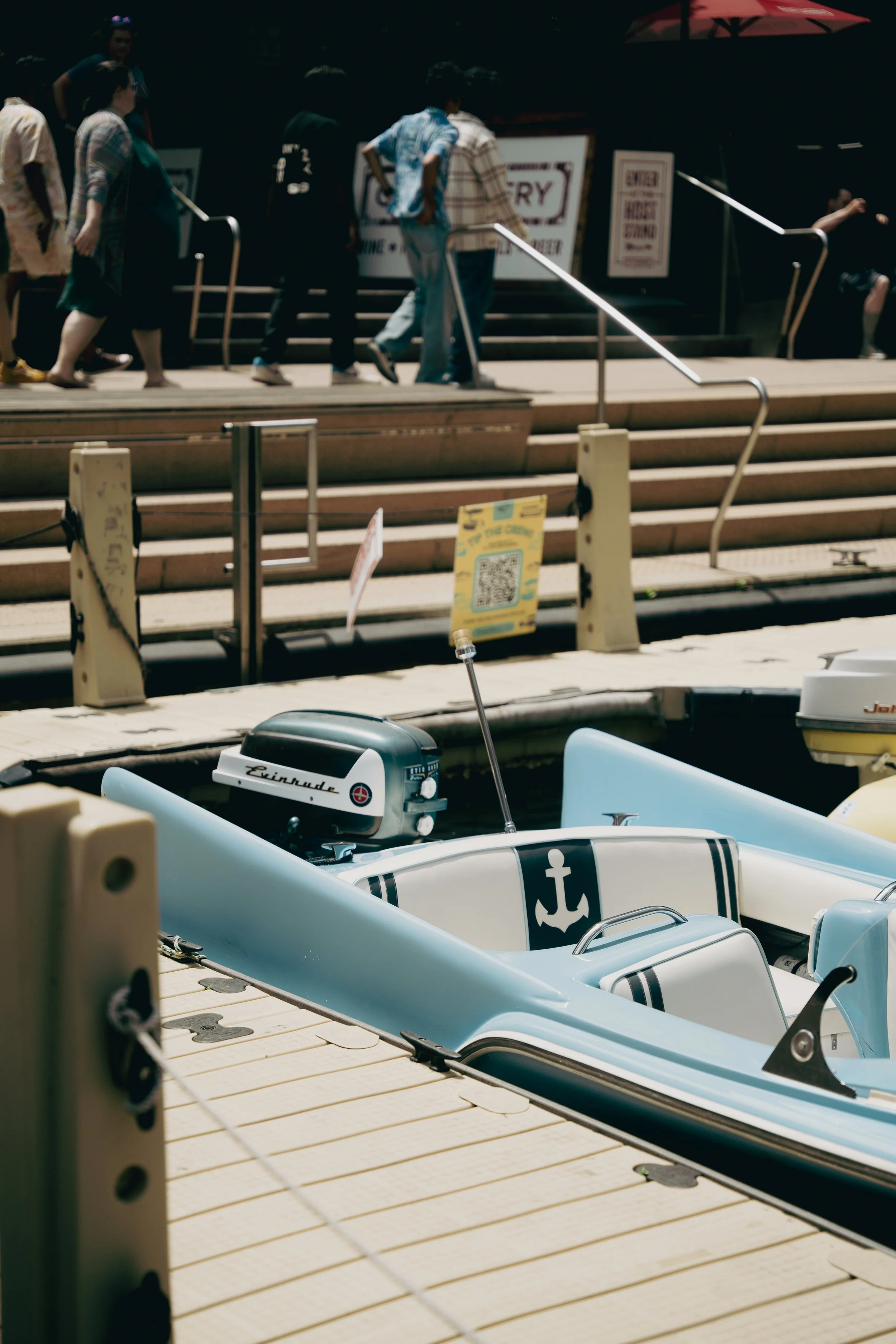 A light blue and white boat with an anchor logo on the seat, docked at a marina, with a wooden walkway and people walking in the background.