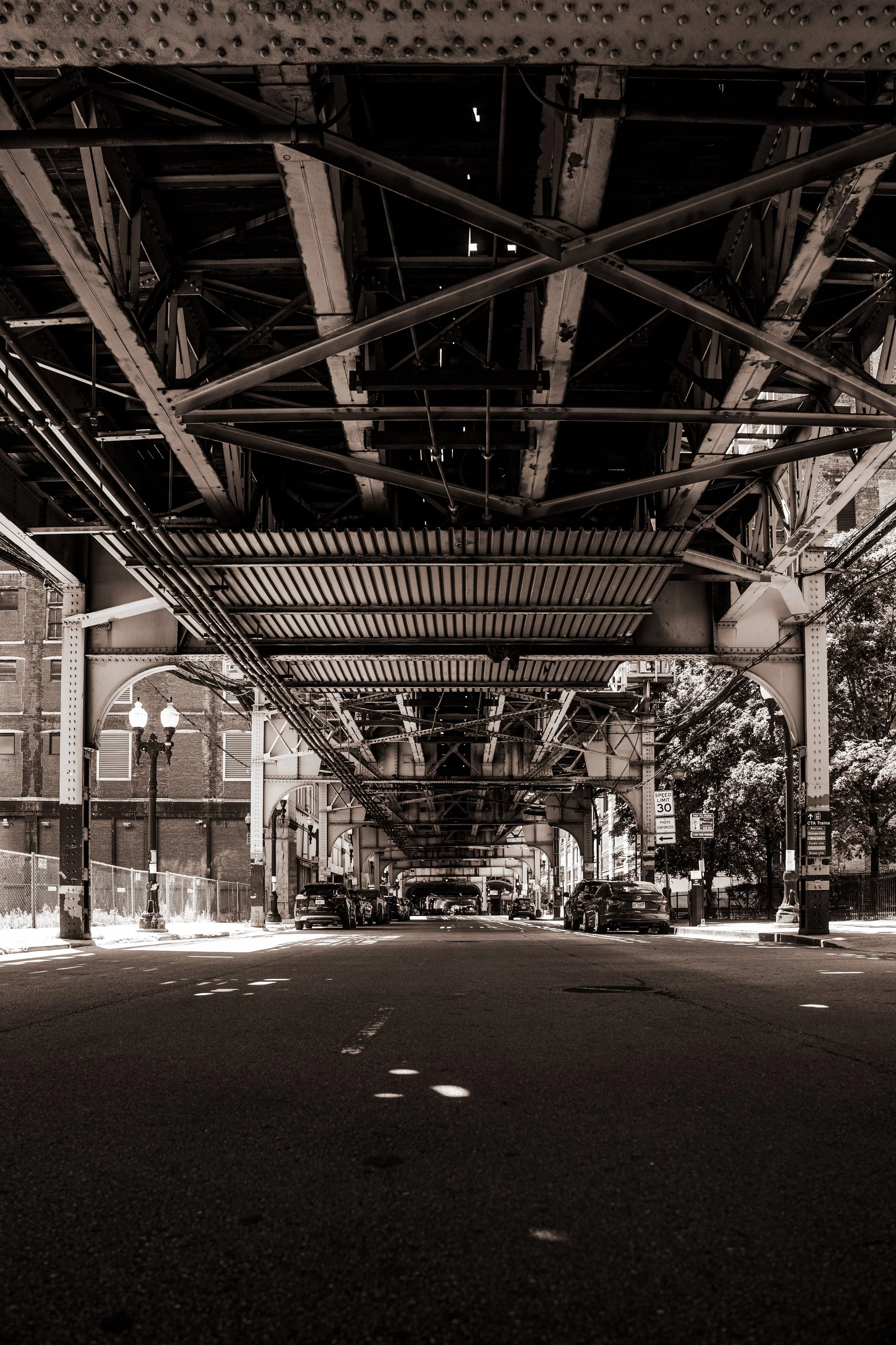 Underneath an elevated train track Chicago's Loop, view of a street with parked cars and street lamps, black and white image.