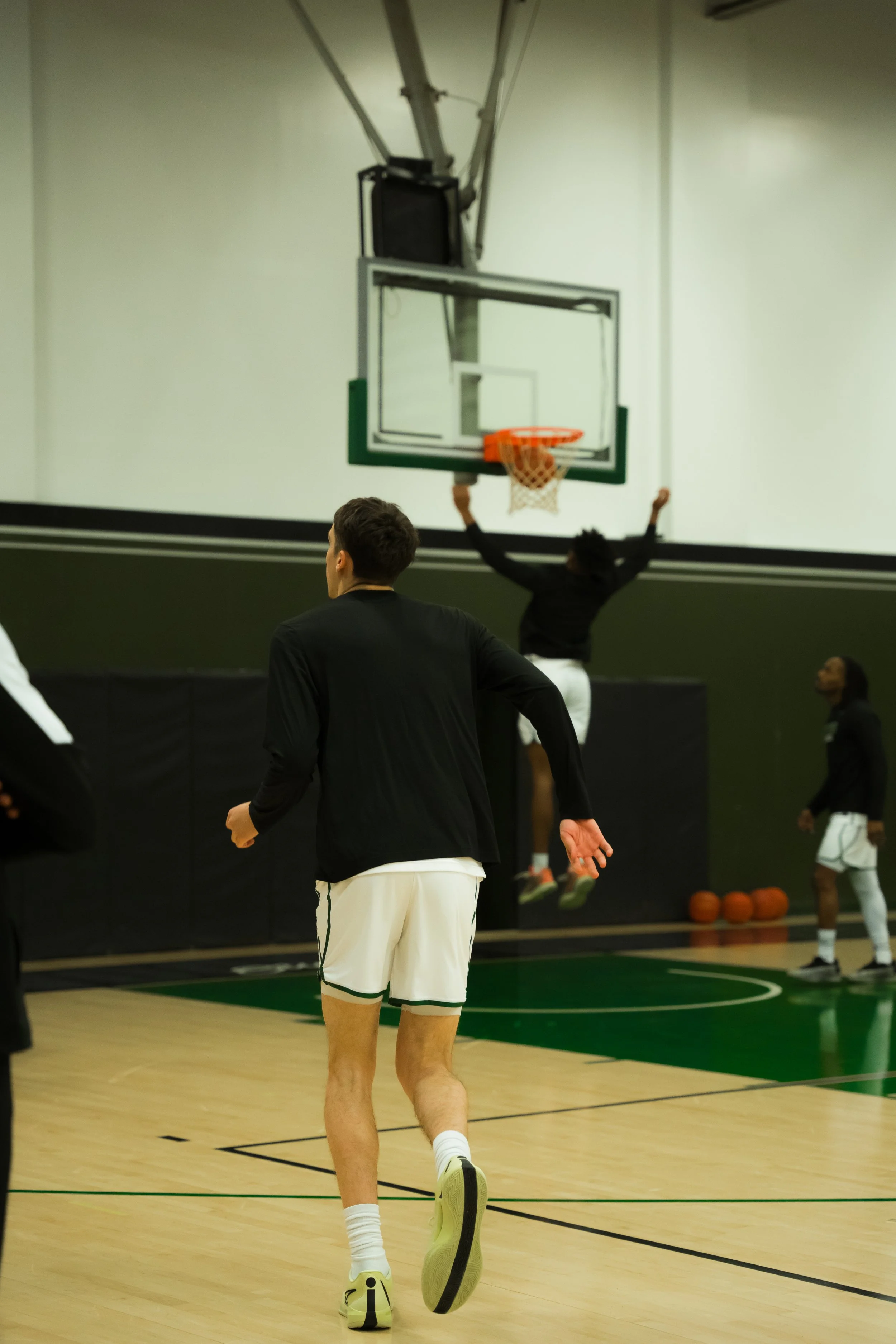 People playing basketball in an indoor gym, with one person jumping near the hoop and another running on the court.
