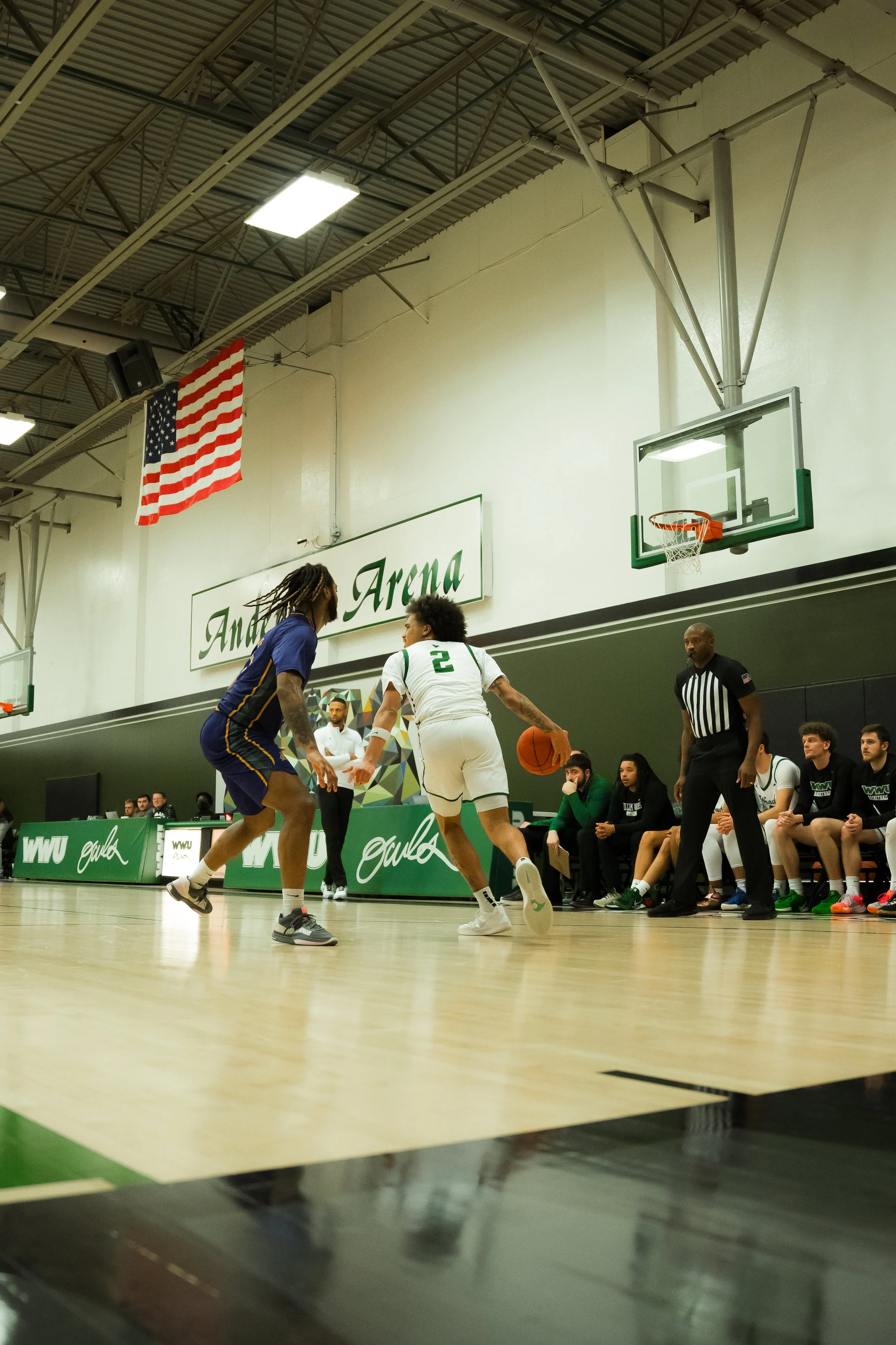 Two basketball players compete for the ball during a game at an indoor gymnasium, with a referee and benches full of players and coaches in the background.