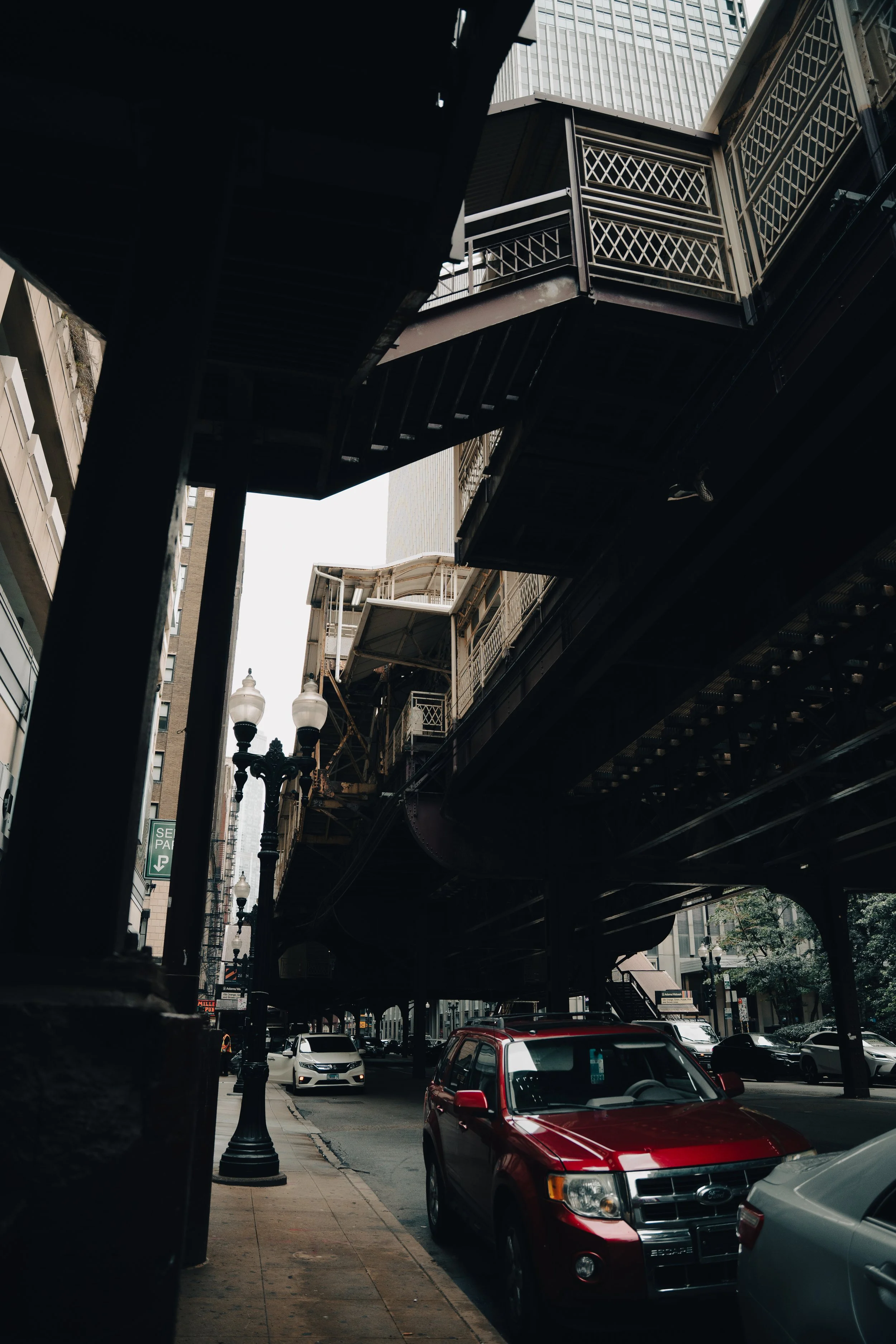 A view of a city street under an elevated train track Chicago's Loop, with cars parked along the curb and a few moving cars in the background, and ornate light posts lining the sidewalk.