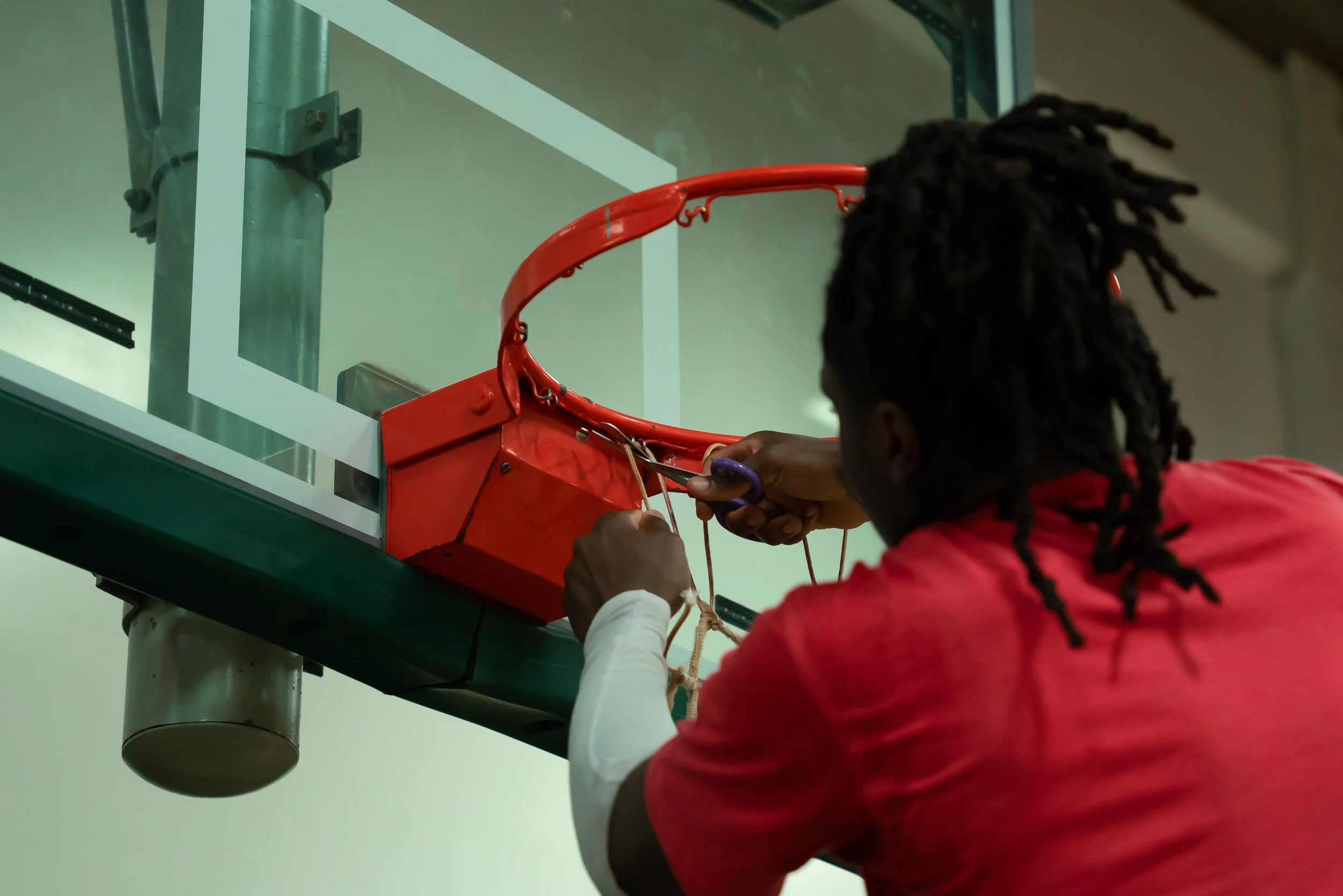 A person with dreadlocks and a red shirt fixing a broken basketball rim.