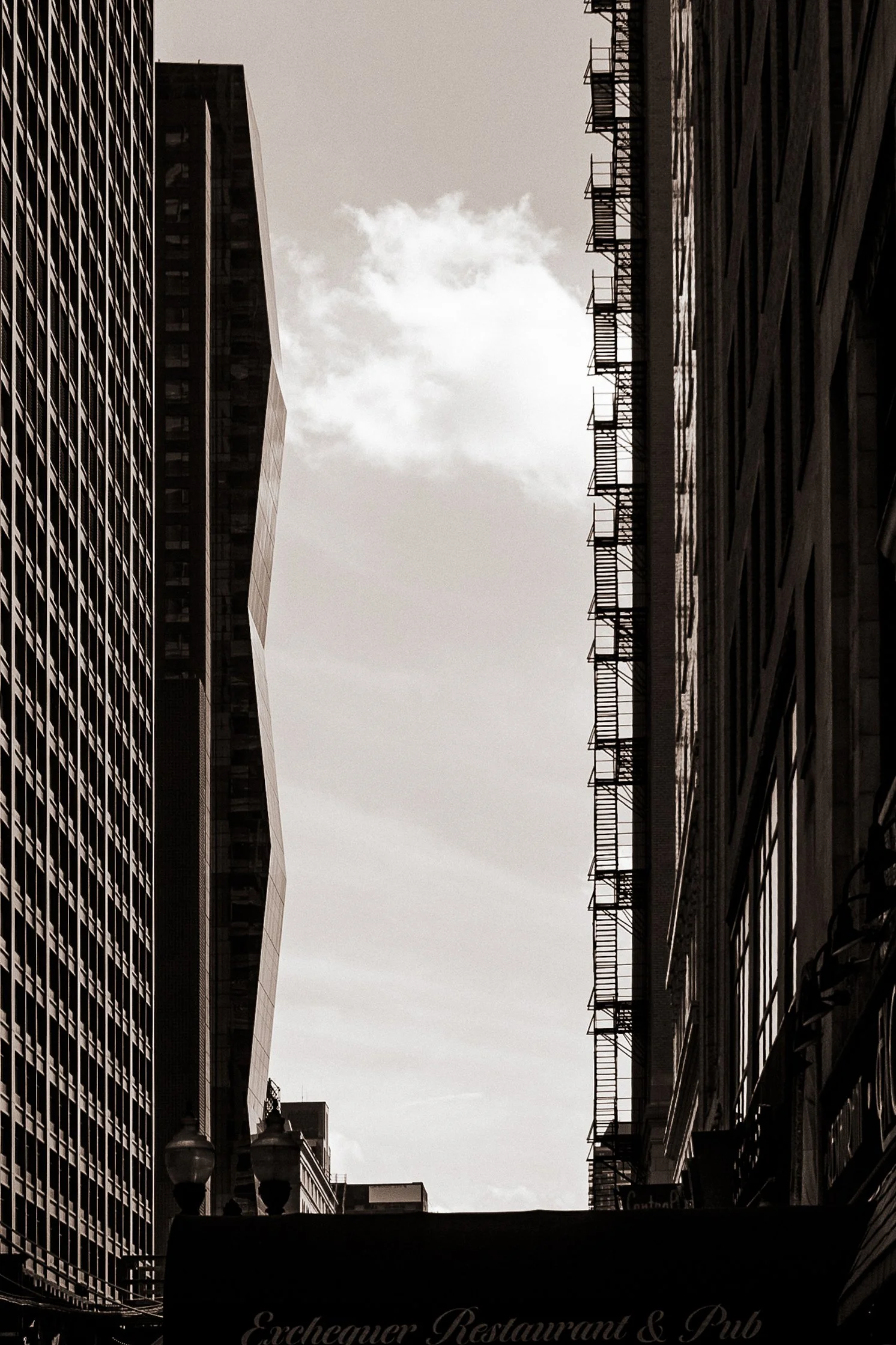 Black and white photograph of tall city buildings with fire escapes on the right, and a building with a distinctive angular design on the left, under a cloudy sky.