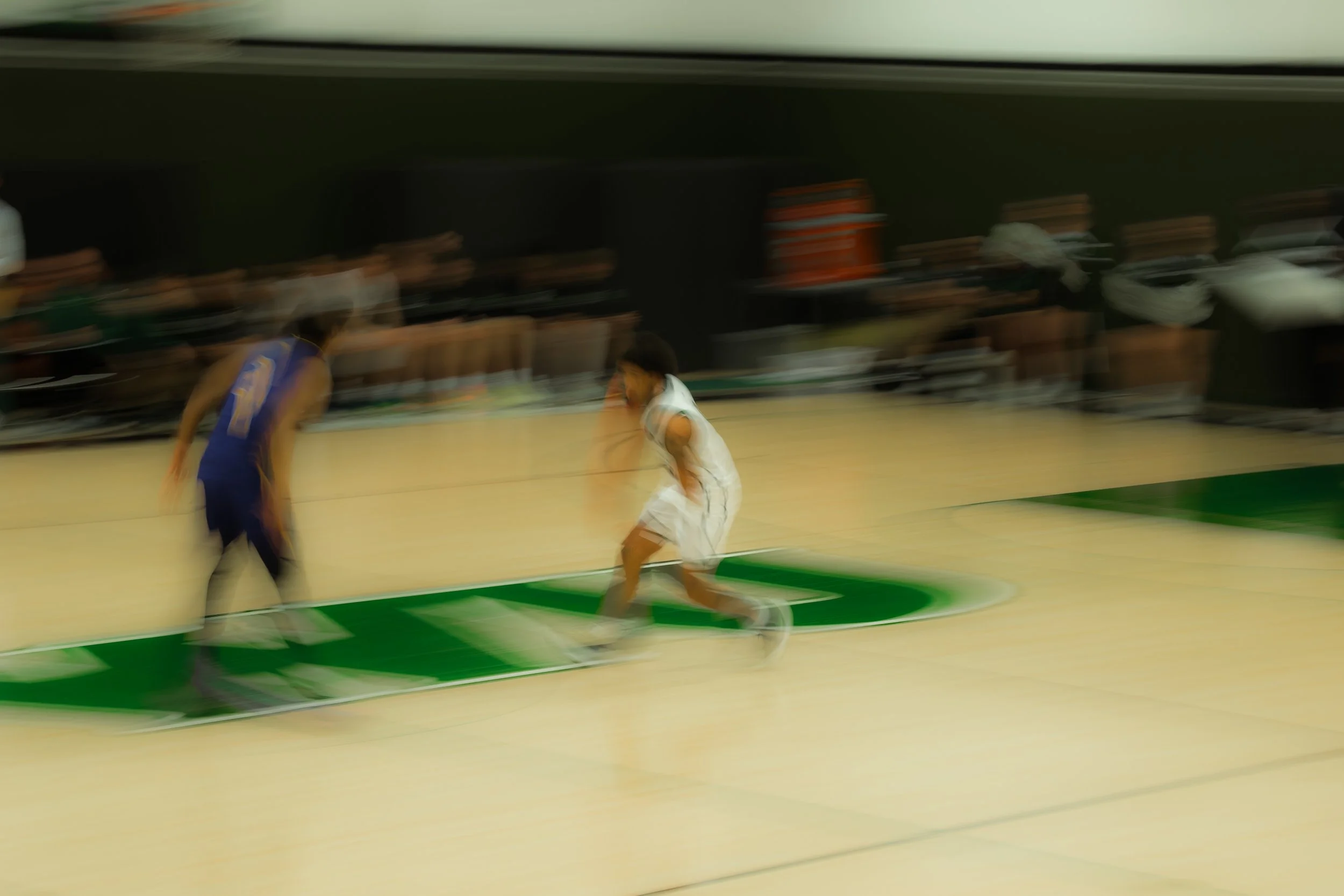 Blurred image of two young basketball players in action on the court, one in a white jersey and the other in a blue jersey, with teammates sitting on the bench in the background.