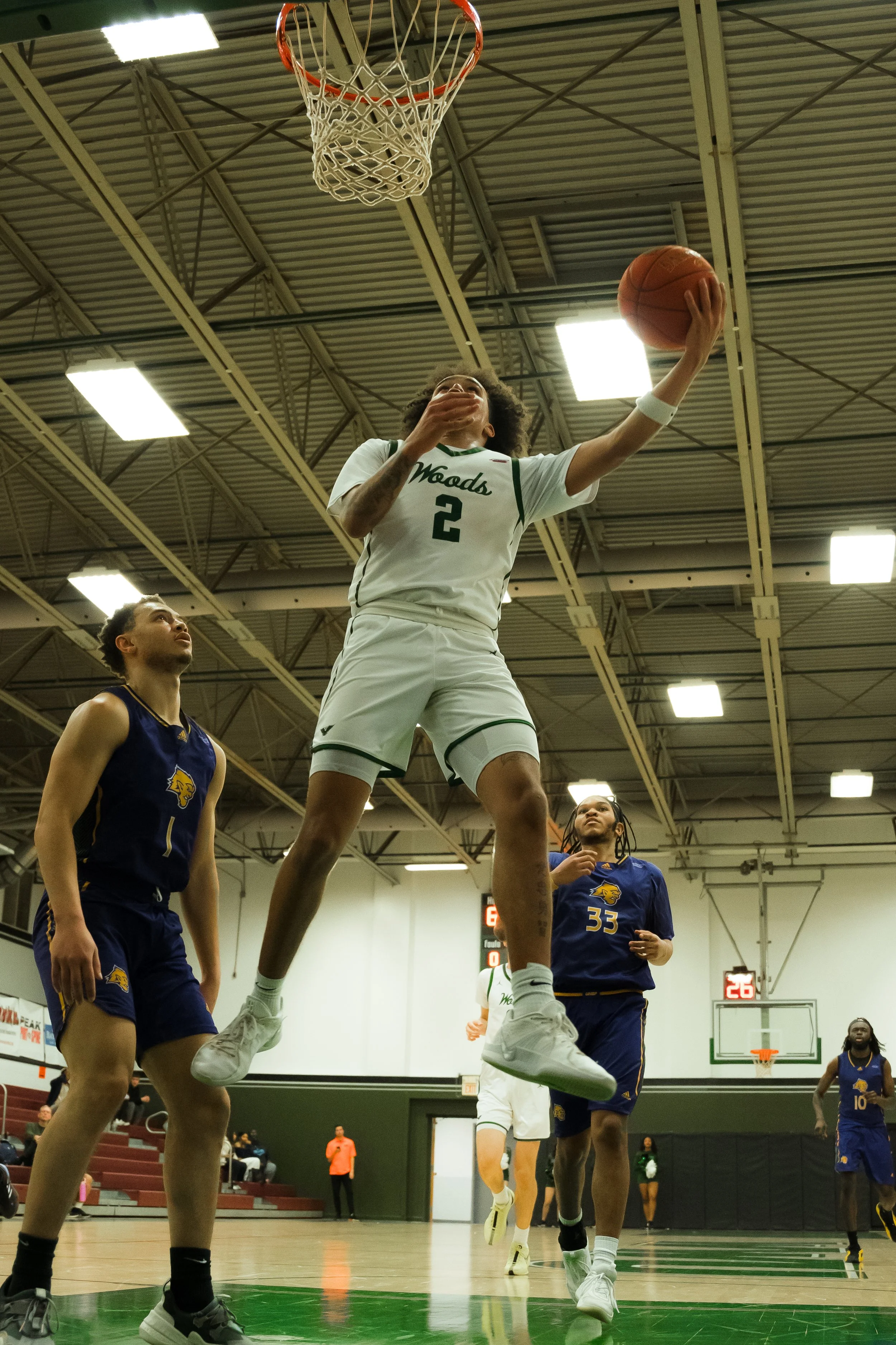 A basketball player in a white uniform with green accents jumping for a layup or dunk, with two opponents in dark blue uniforms attempting to defend, all inside an indoor basketball court.