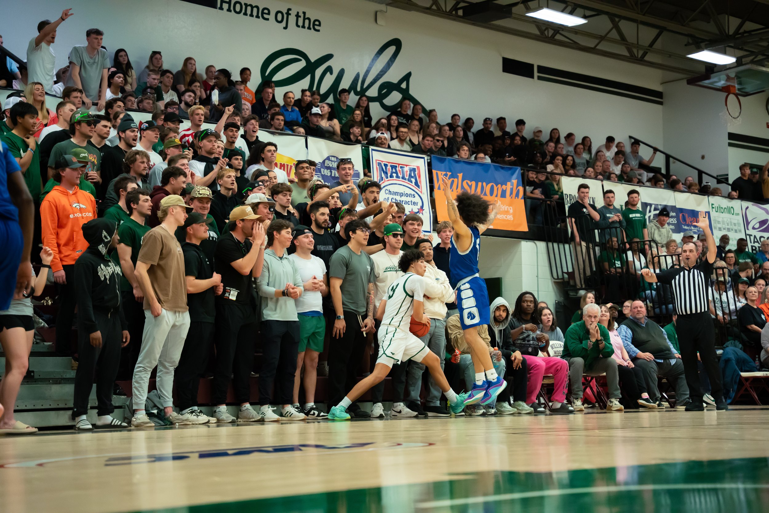 A basketball game in a gymnasium with a crowd, where a player in a white uniform is jumping for the ball against a player in a blue uniform, while a referee is raising his arm for a call.