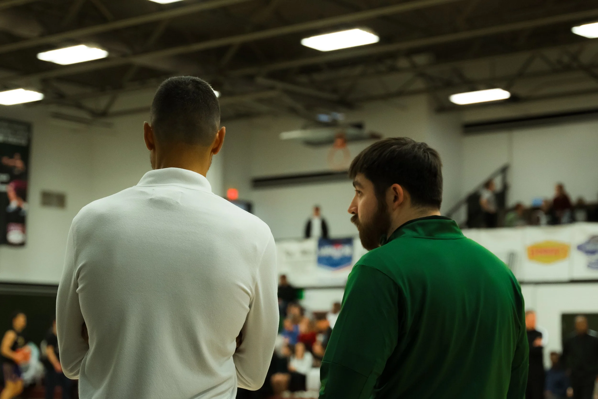 Two men talking in a gymnasium, with people sitting and standing in the background.