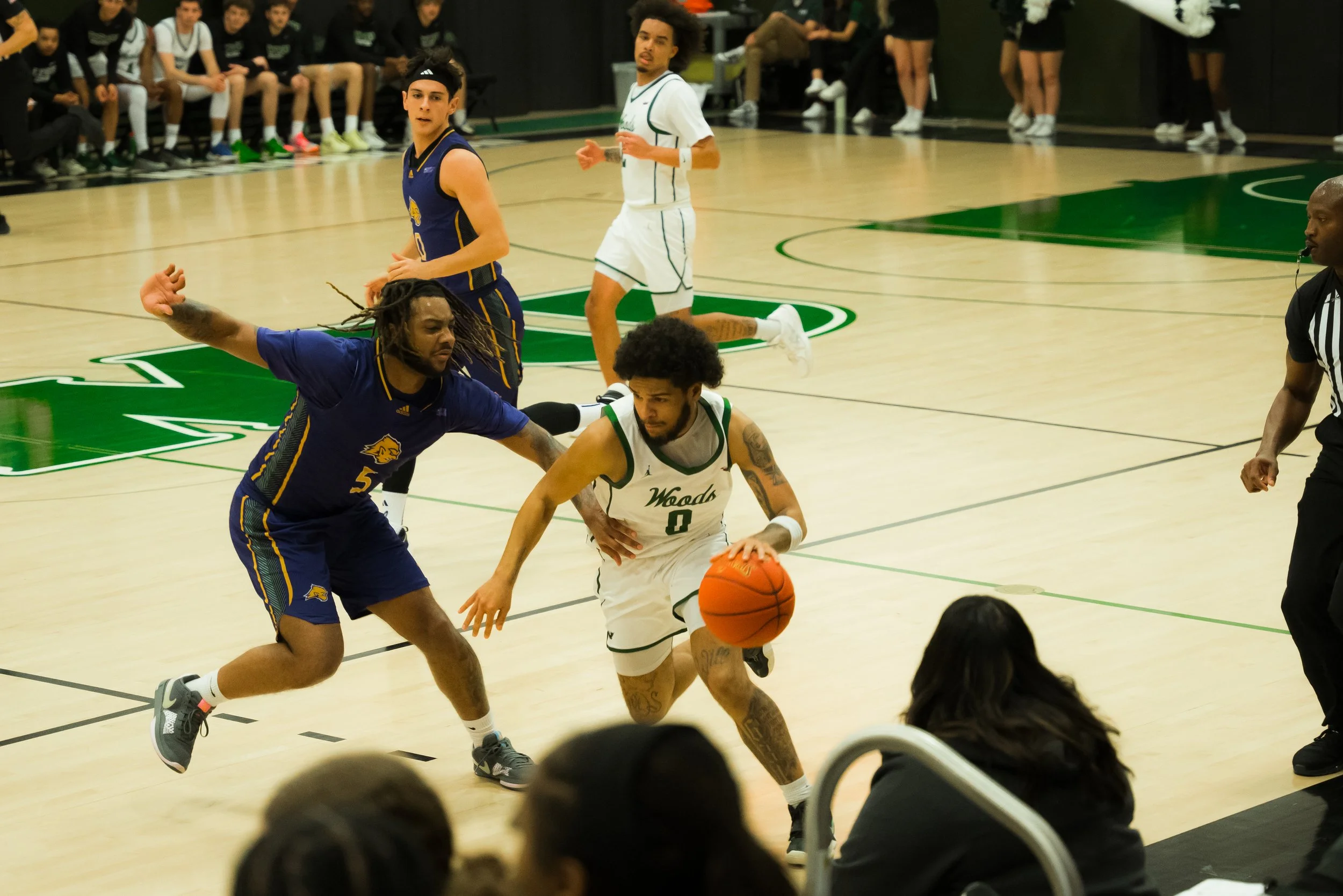 Two basketball players compete for possession of the ball during a game, with other players and spectators in the background.