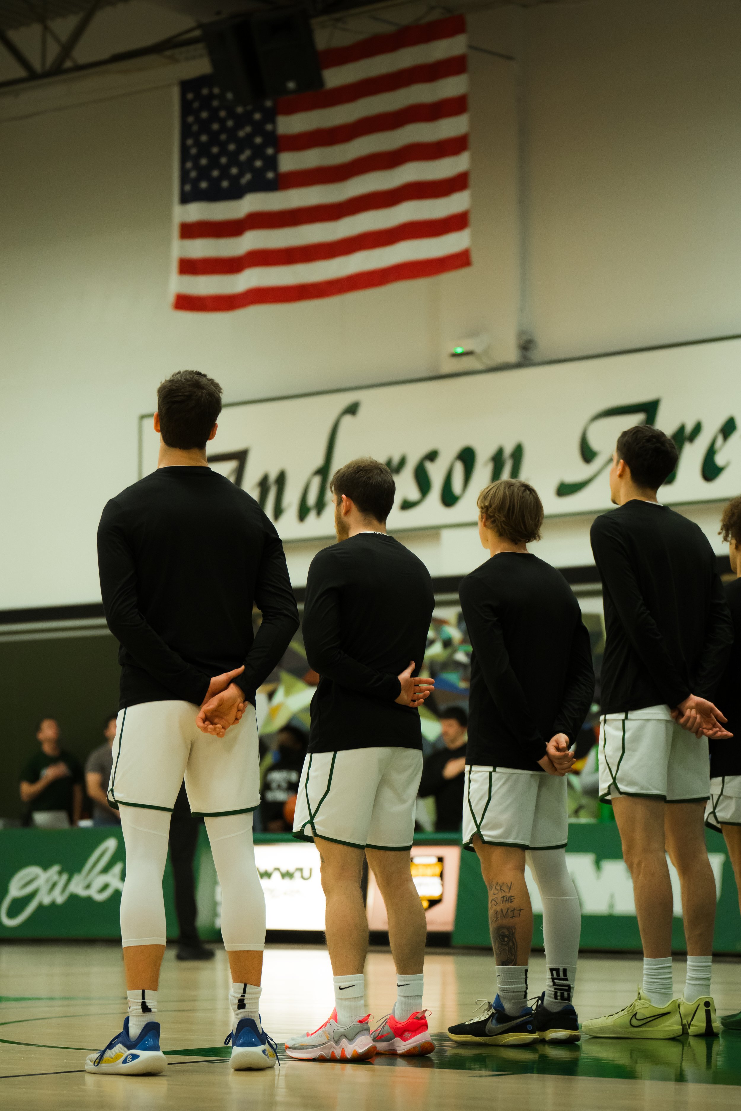 A group of basketball players standing on a court, facing away from the camera. They are dressed in black tops and white shorts, with various colorful athletic shoes. An American flag hangs on the wall behind them, and a sign reads 'Underson.'