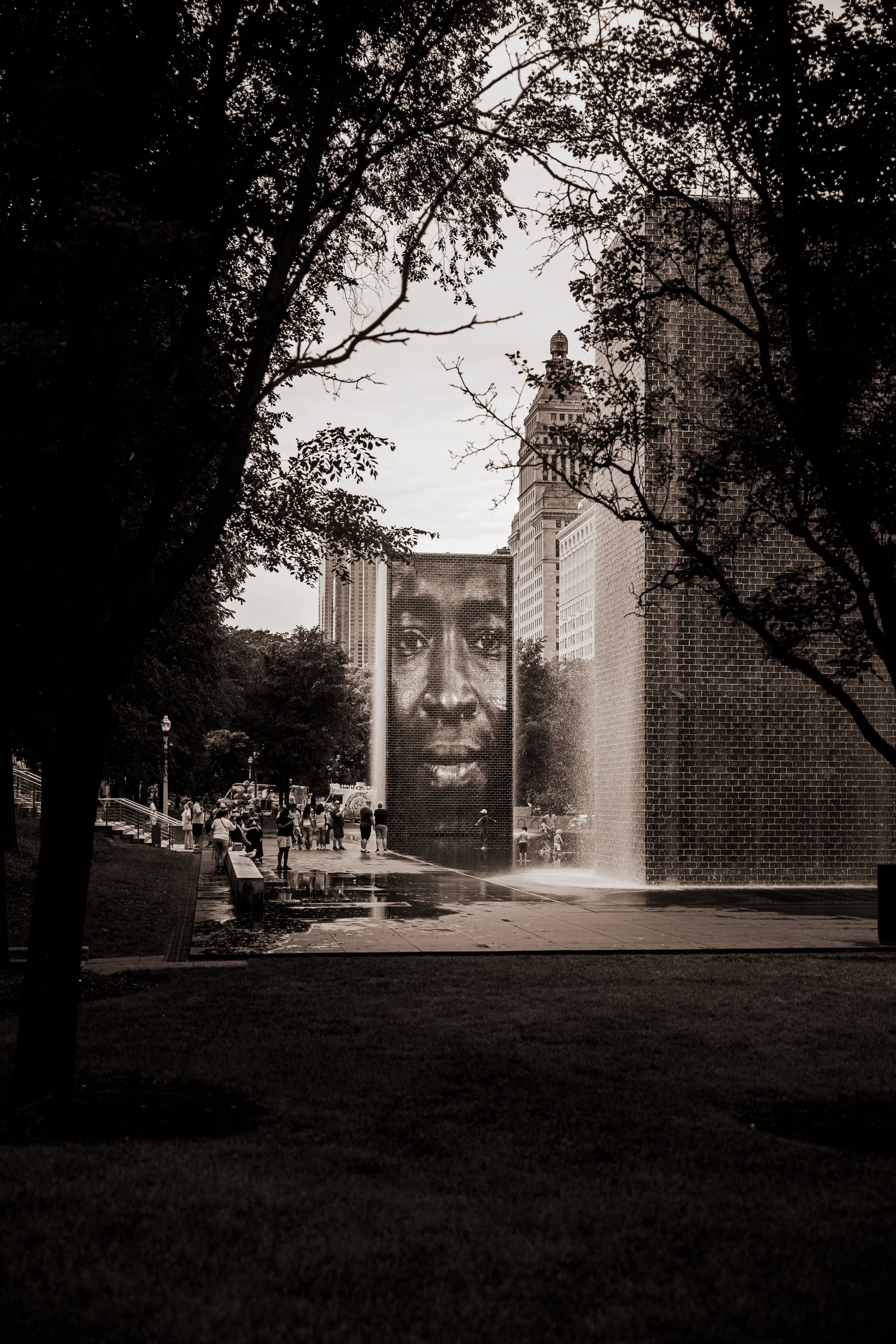 Fountain with a large digital screen displaying a woman's face, surrounded by trees and a group of people at a park.
