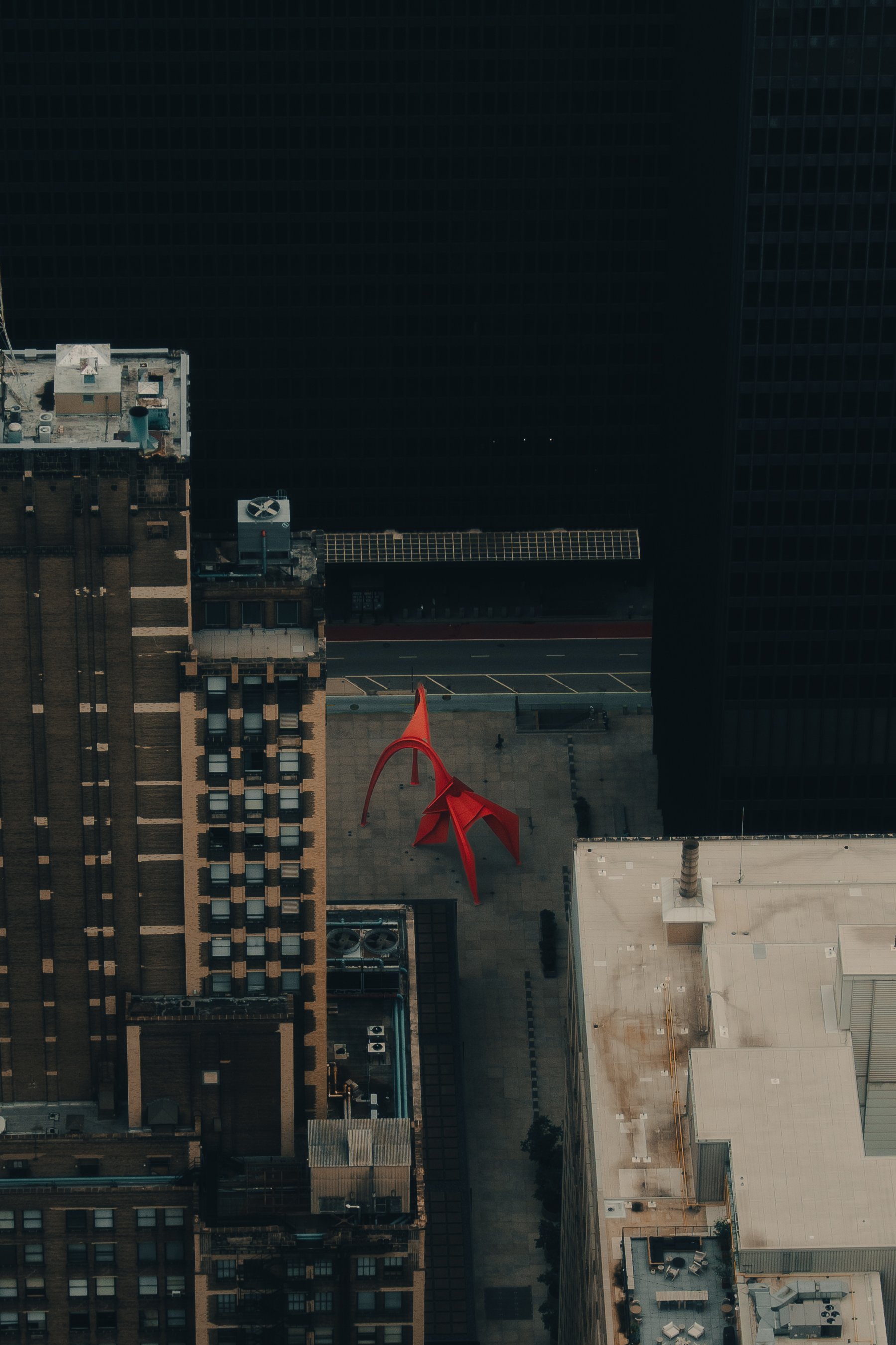 Aerial view of a city rooftop with a large red sculpture on a concrete plaza, surrounded by tall buildings and dark skyscrapers.