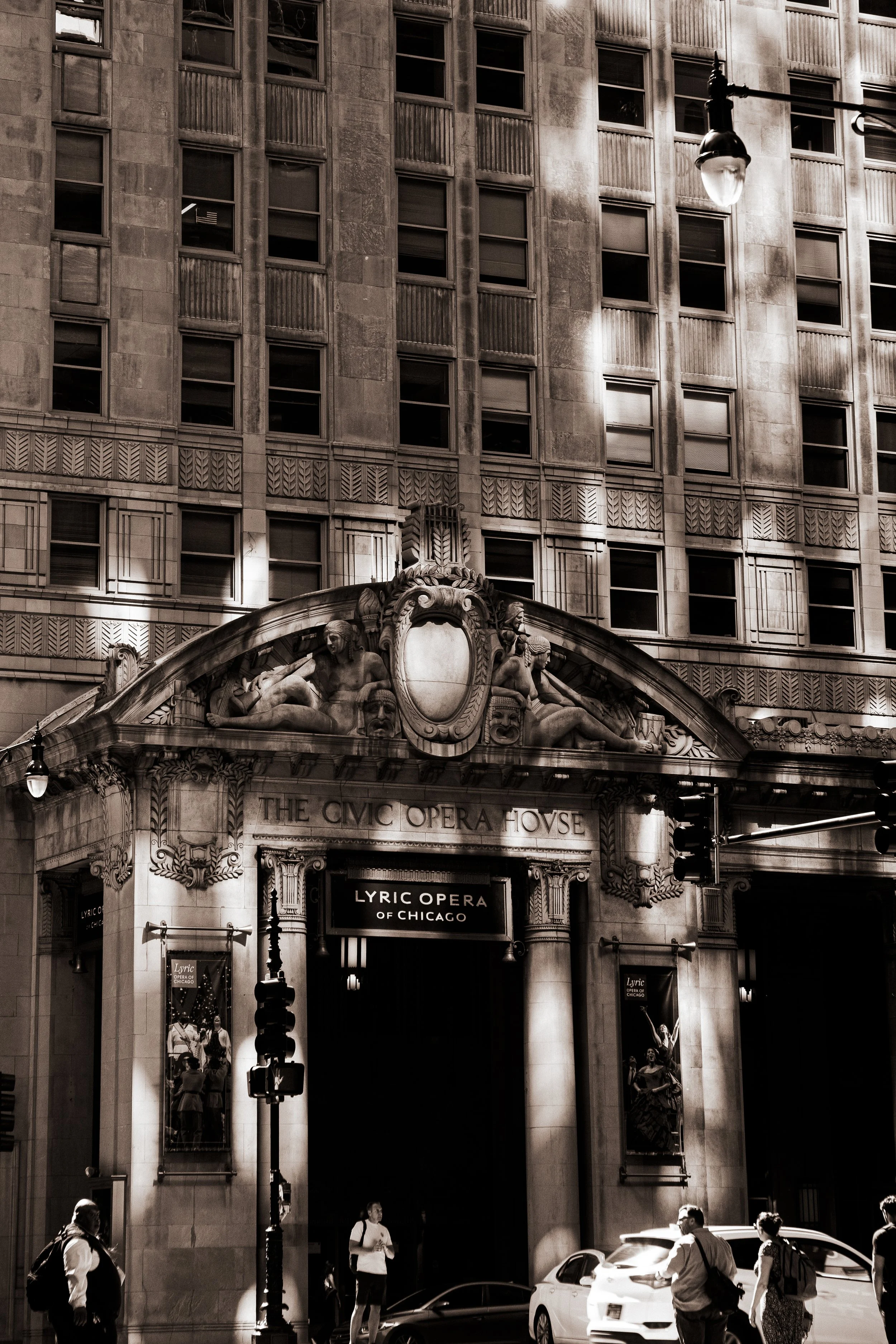 Daytime view of the Lyric Opera of Chicago building with decorative sculptures and lighting, with people and cars in the foreground.