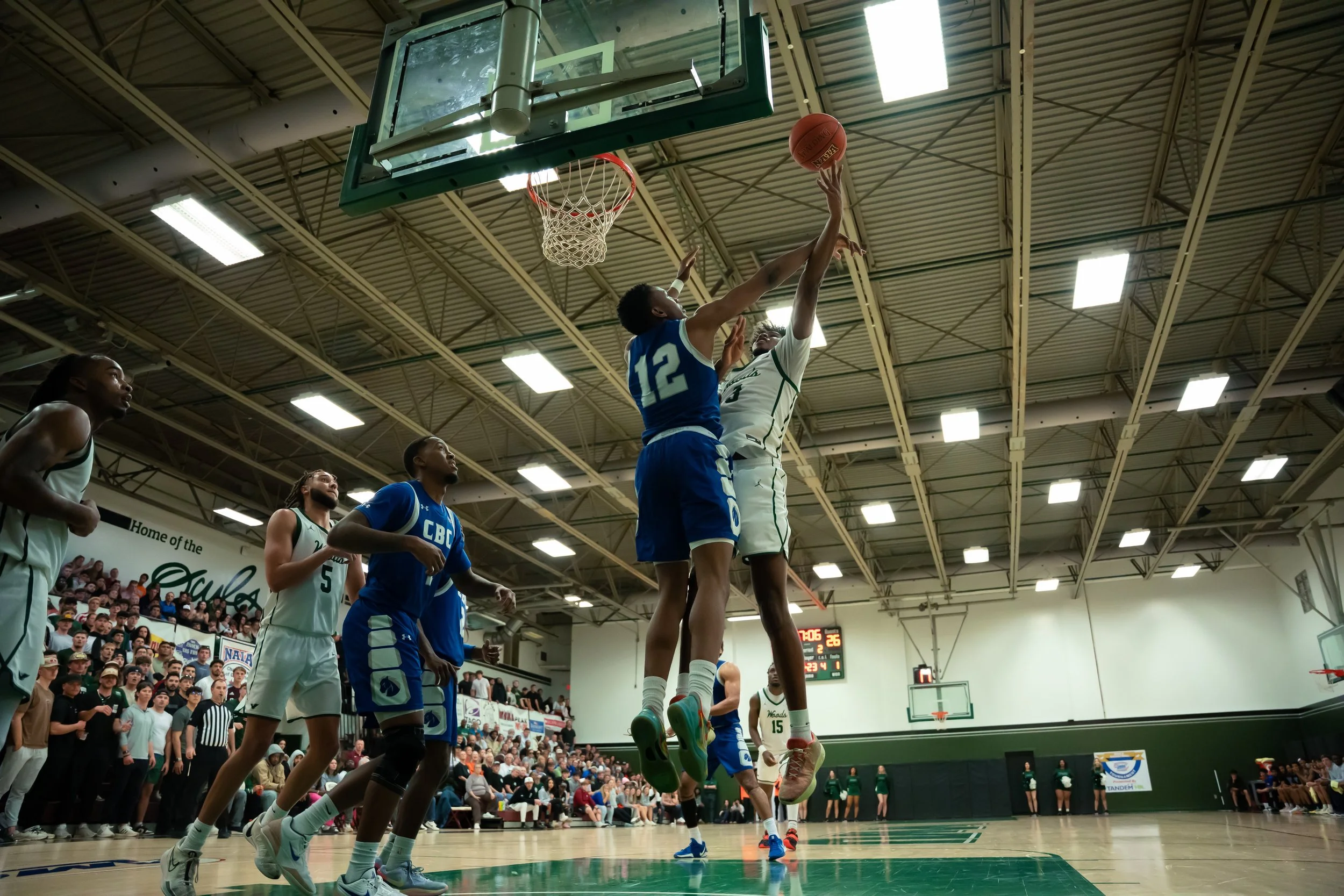 Two basketball players, one in a white jersey and the other in a blue jersey, jump near the basket to contest a shot during a game. Several other players and spectators are visible in the background inside a gymnasium.