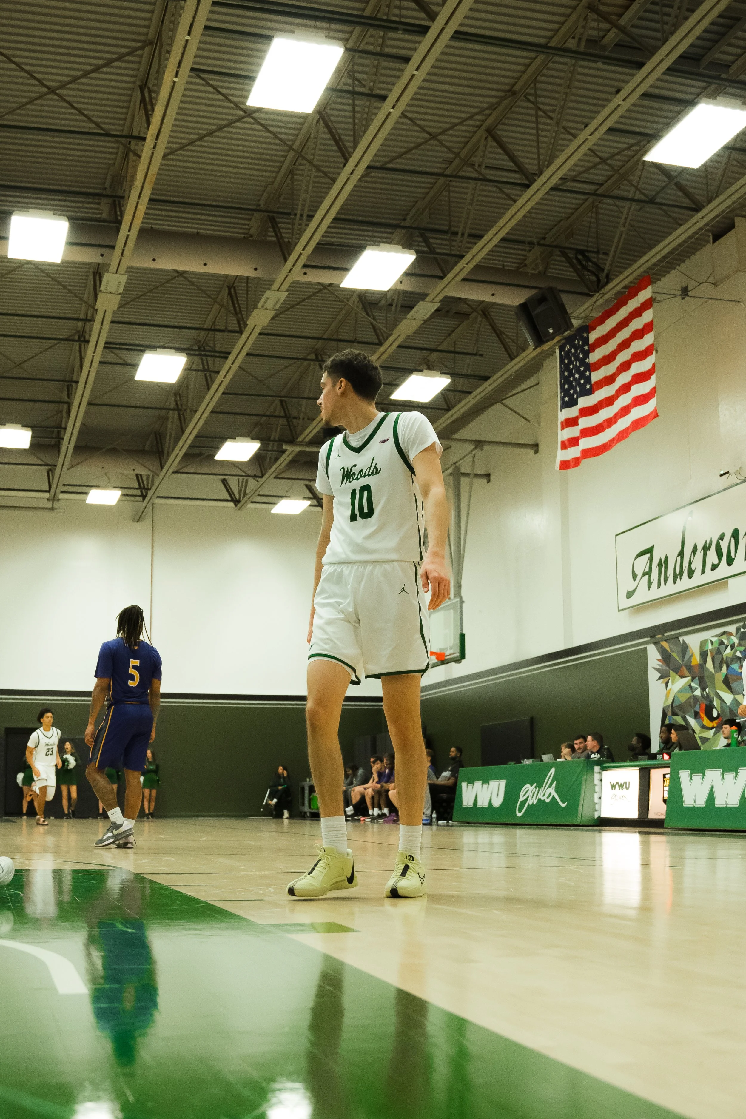 A basketball player in a white jersey with green accents, number 10, standing on a basketball court. Other players and spectators are visible in the background, with an American flag hanging on the wall.