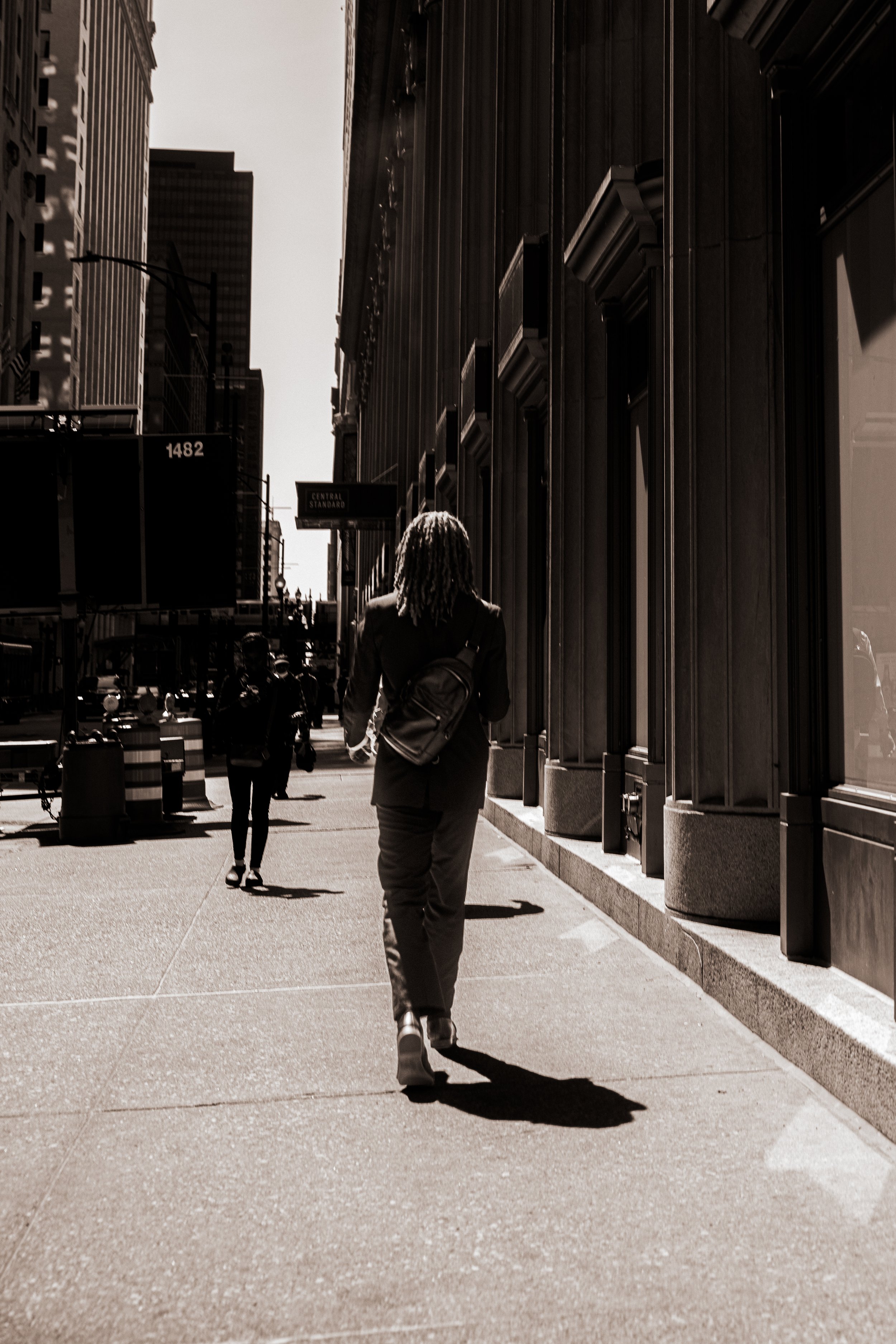 A black-and-white photo of a woman walking on a city sidewalk, carrying a backpack, with tall buildings along the street.