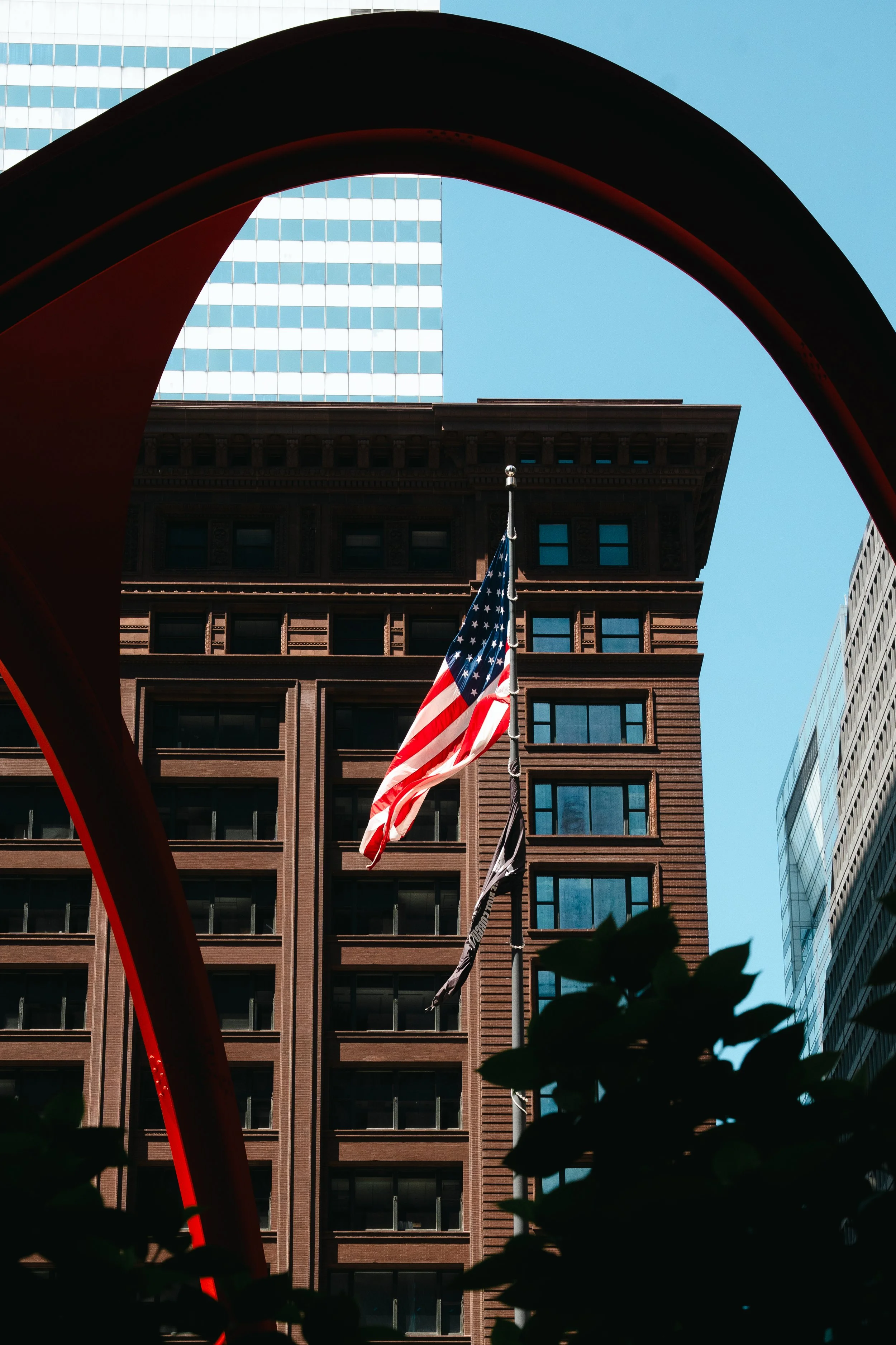 A view of the American flag flying outdoors in front of a tall building, framed by a large, red, curved architectural element. The background features modern office buildings with glass and brick facades and a clear blue sky.