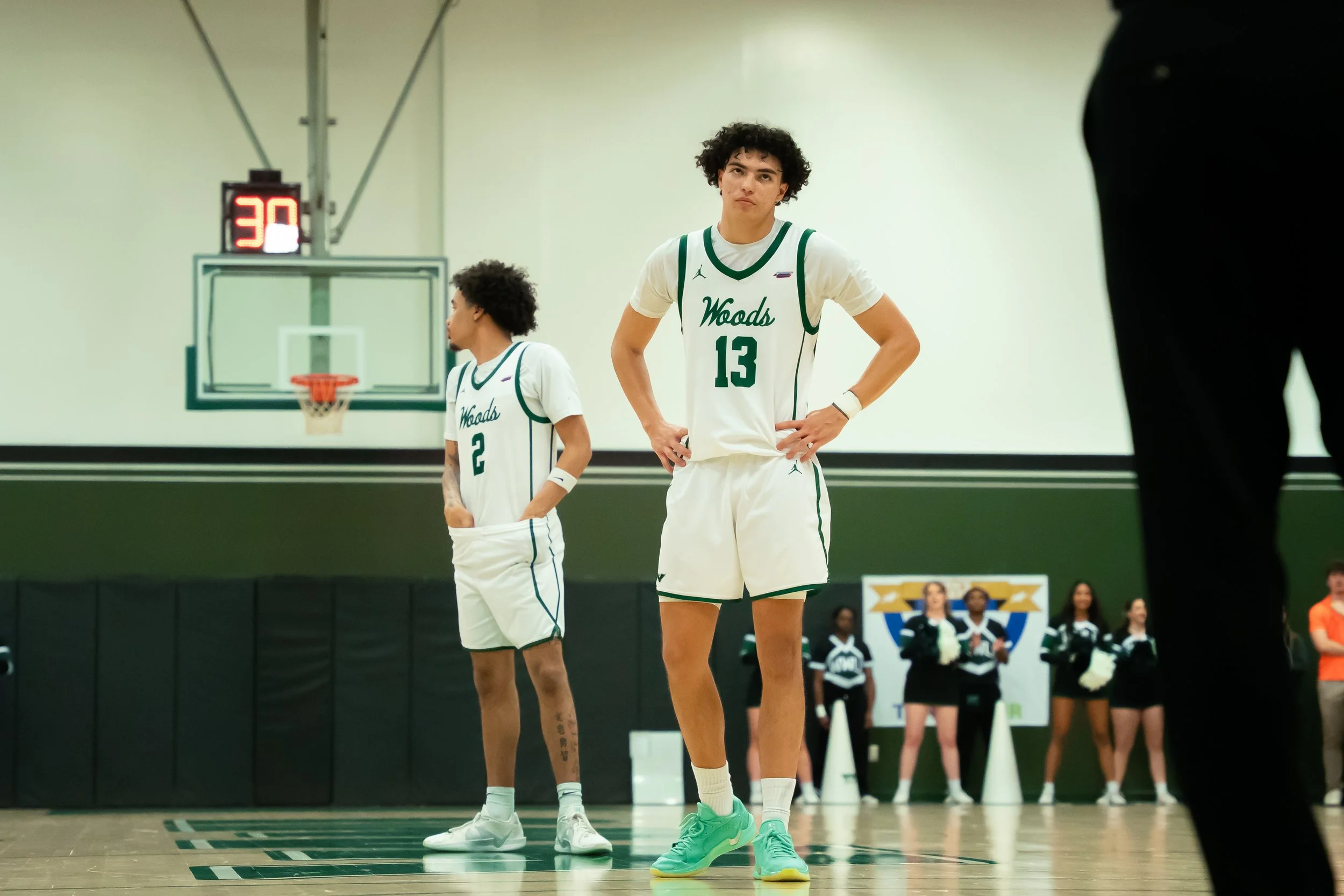 Two basketball players in white and green uniforms standing on the court during a game or practice, with cheerleaders and a referee visible in the background.