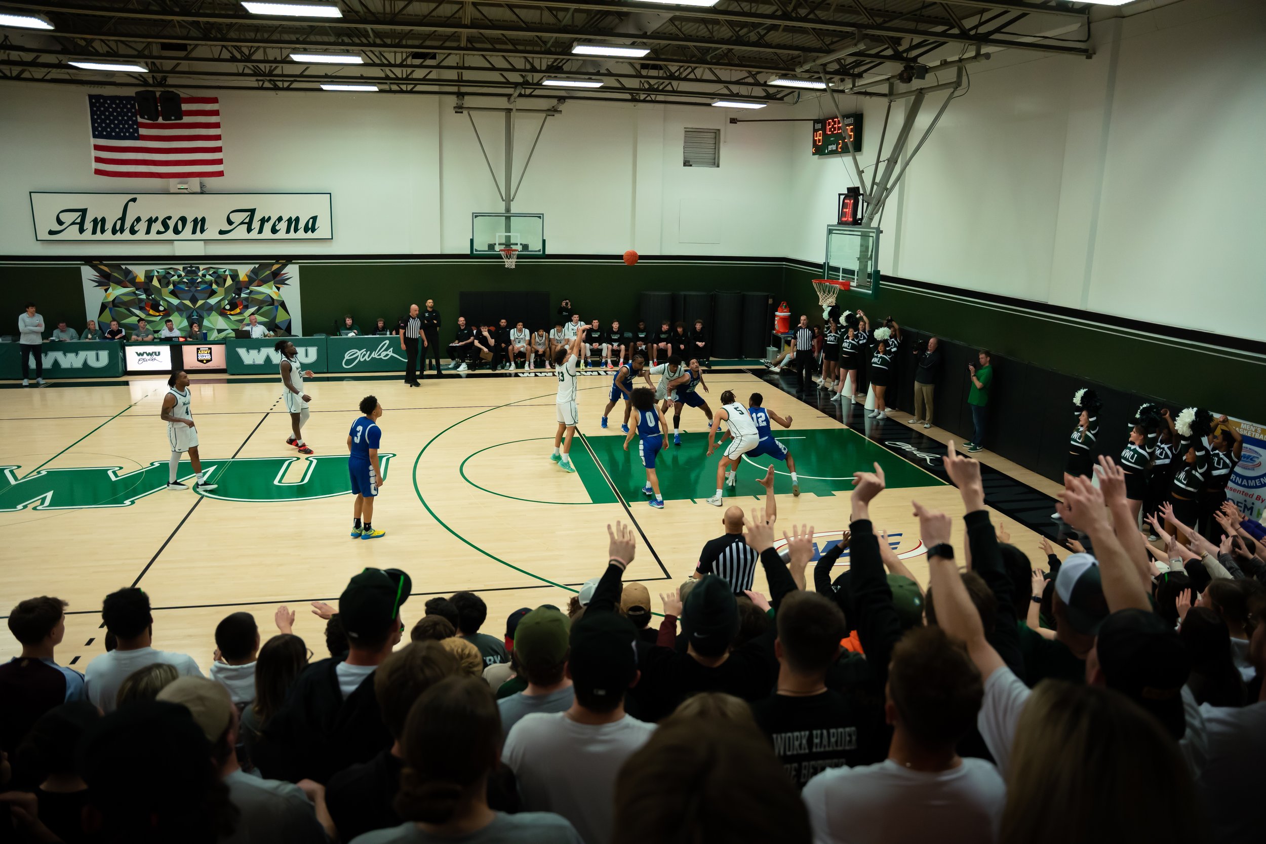 An indoor basketball game at Anderson Arena with players mostly in white and blue uniforms. Spectators cheer in the foreground, and cheerleaders stand along the sidelines. The court features green and white markings, and the scoreboard shows 49-42 wi