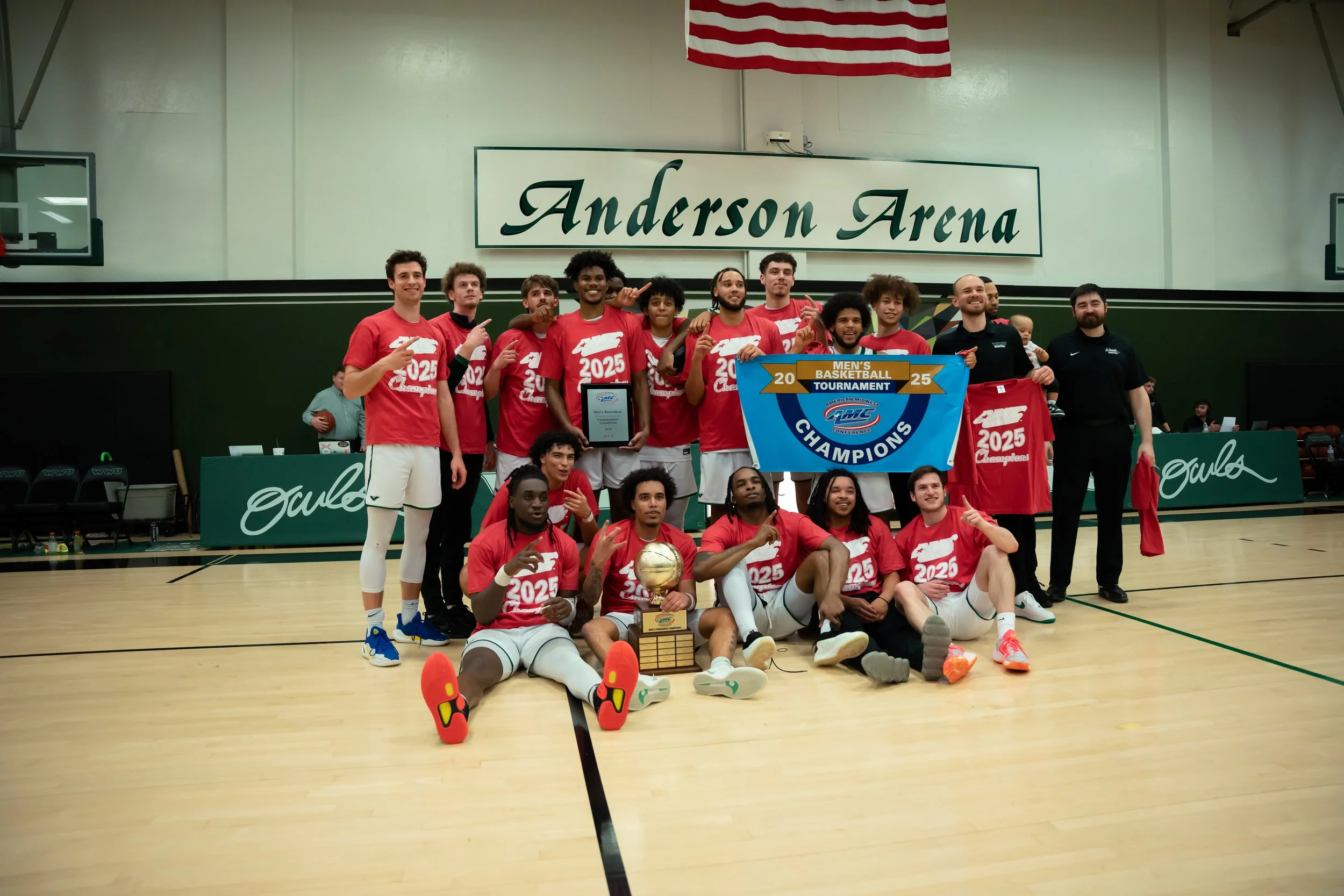 A basketball team celebrating their championship victory at Anderson Arena, holding a blue banner and a trophy, with players and coaches posing on the court.