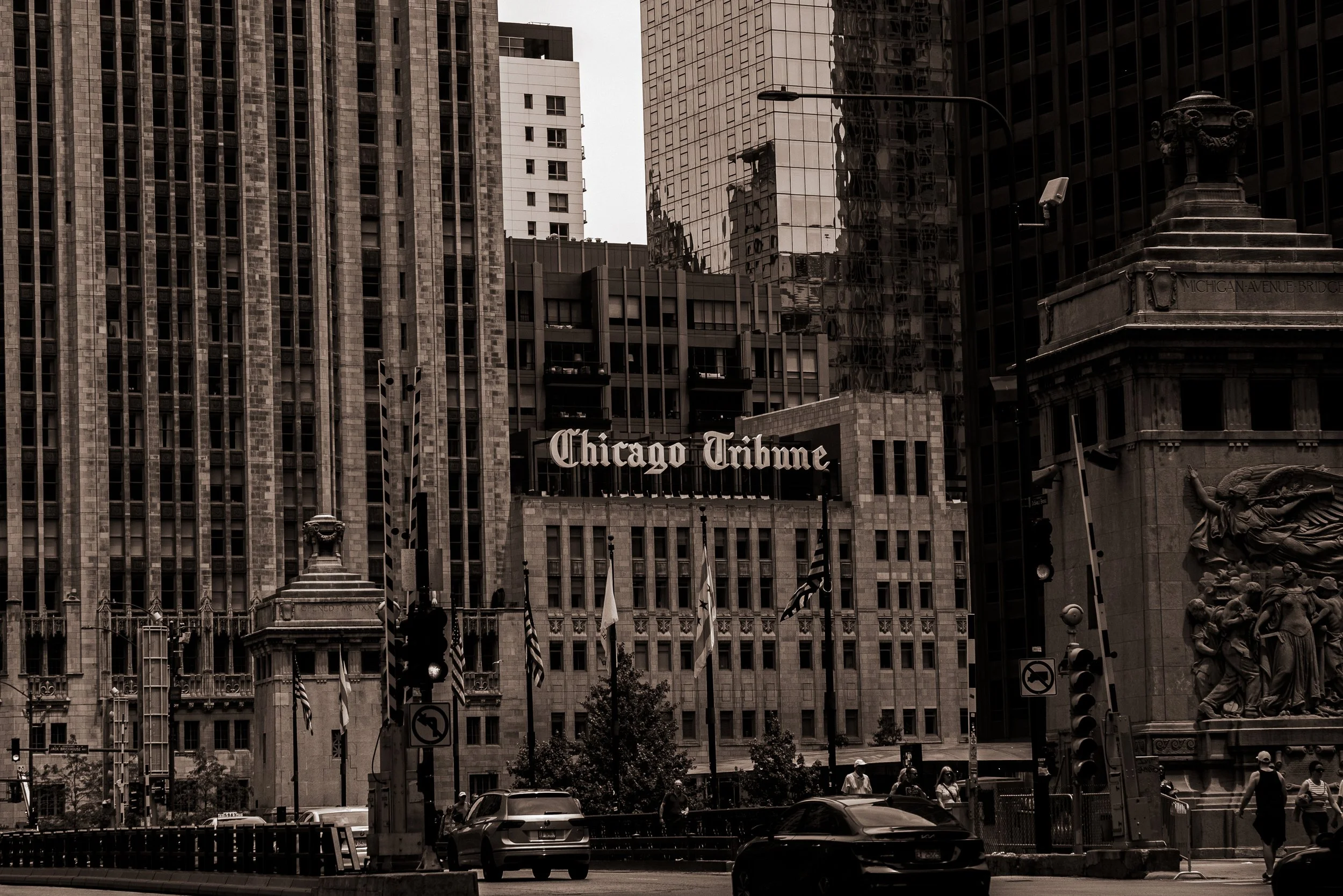 Black and white photo of Chicago Tribune building in downtown Chicago with tall skyscrapers, American flags, and pedestrians.