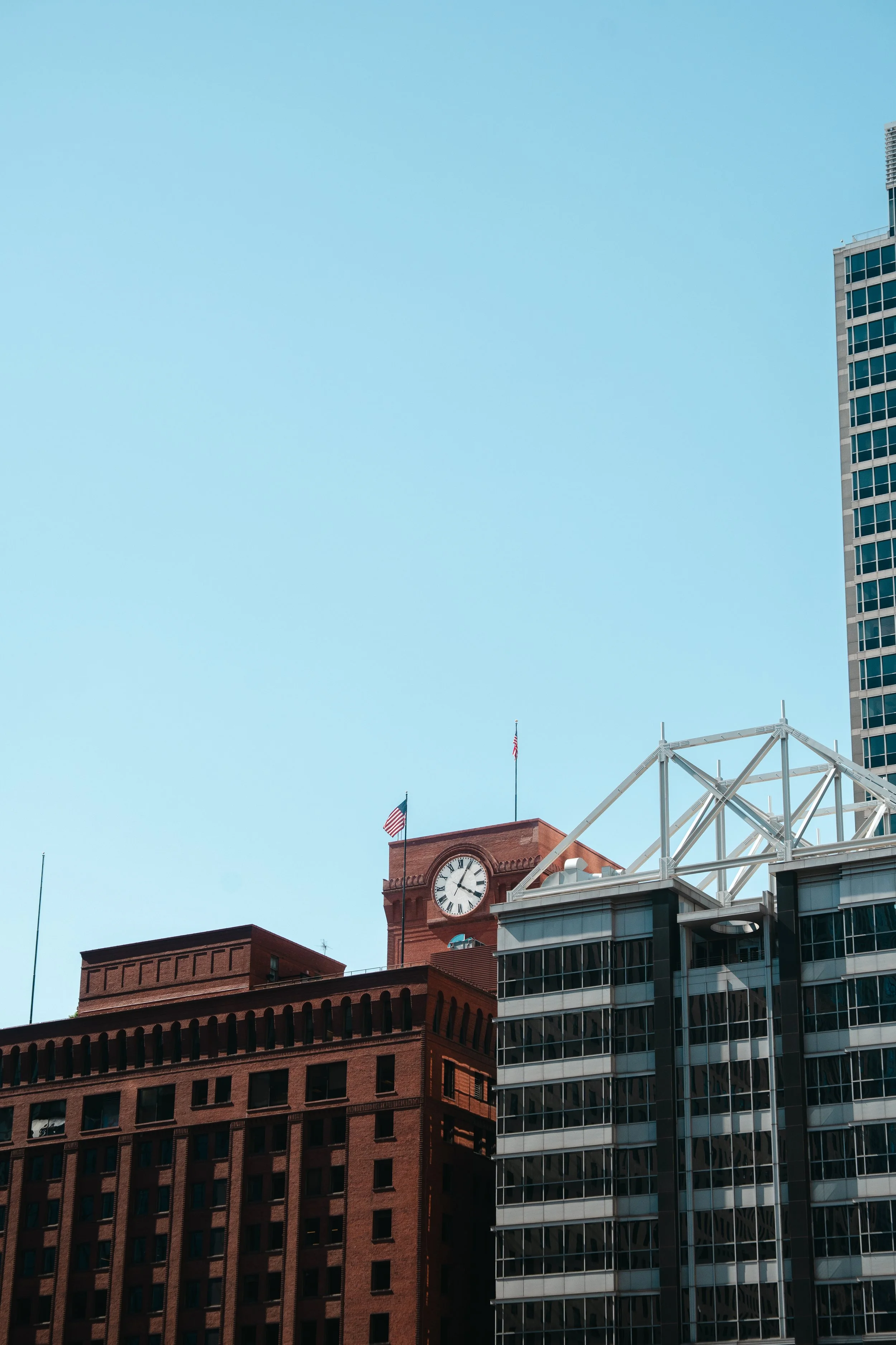 A cityscape featuring a red brick building with a clock tower and American flags, next to modern glass and steel skyscrapers, under a clear blue sky.