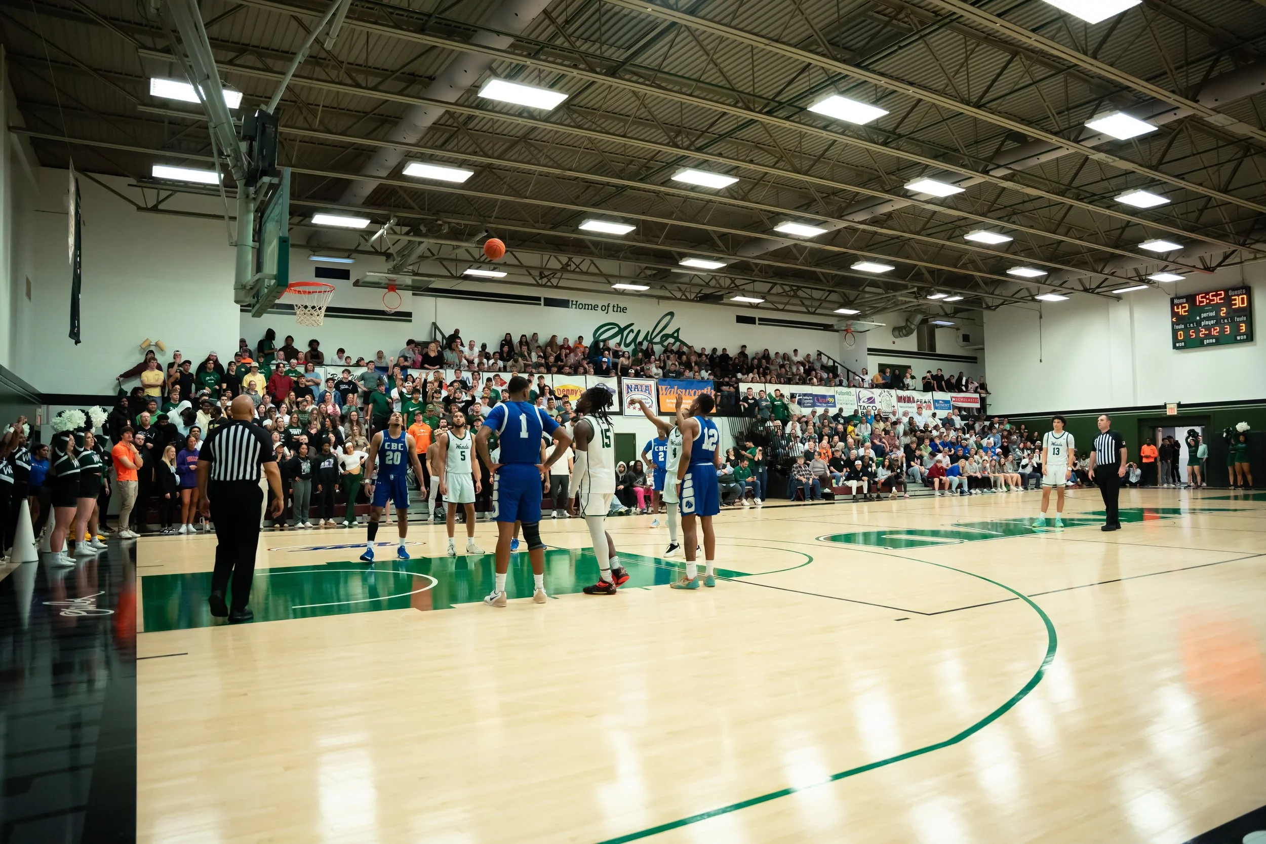 A basketball game in progress with players on the court, referees, and a crowd of spectators in the stands.