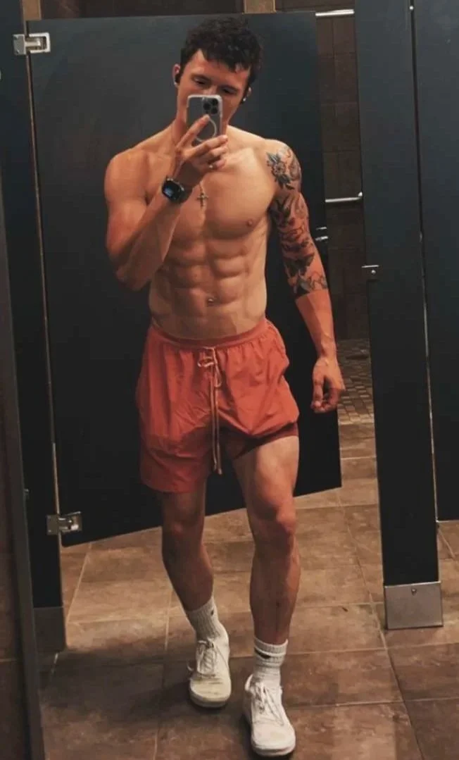 A shirtless young man taking a mirror selfie in a bathroom. He is wearing orange shorts, white sneakers, and socks, with tattoos visible on his left arm and chest.