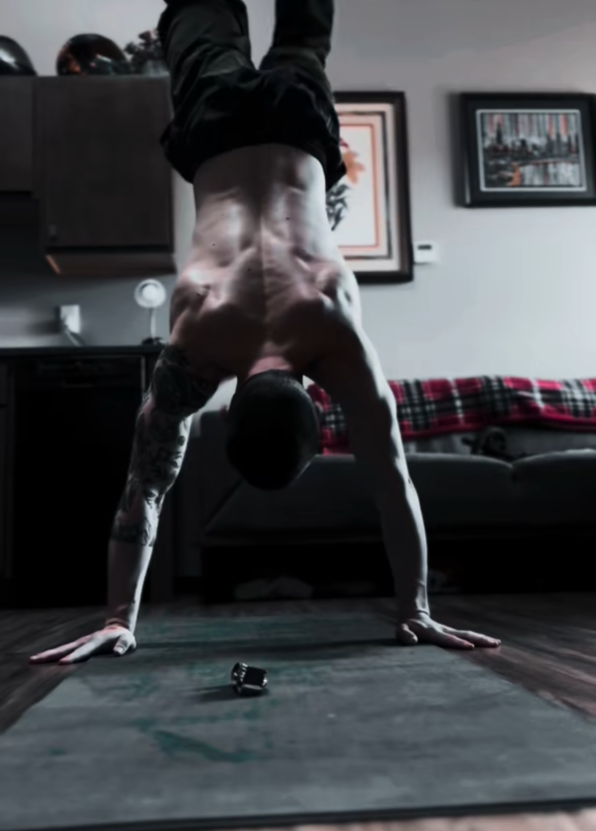 A shirtless man with tattoos performing a handstand indoors, looking down at the camera, in a living room with framed pictures and a plaid blanket on a couch.