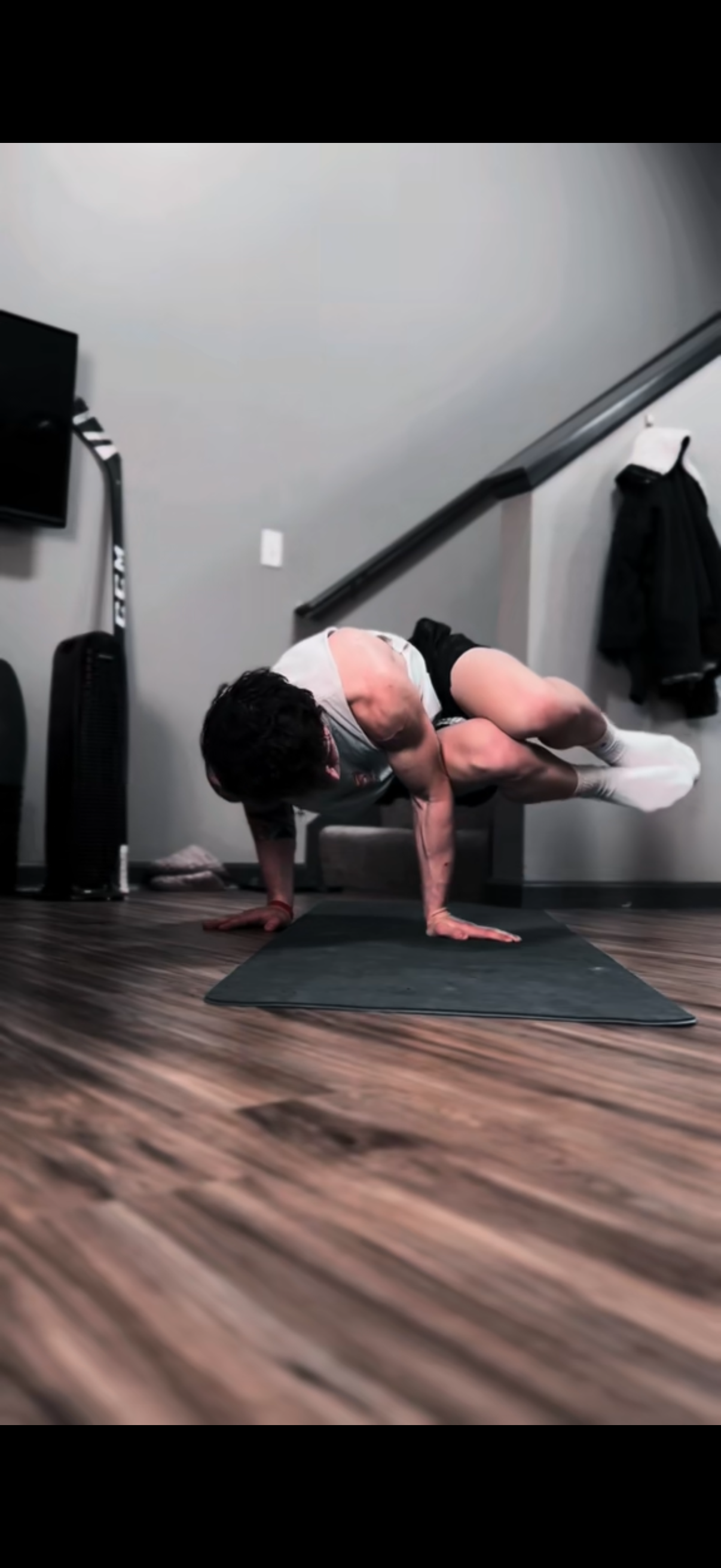 A person performing a challenging yoga pose on a mat in a room with wooden floors and gray walls, with a staircase and television visible in the background.