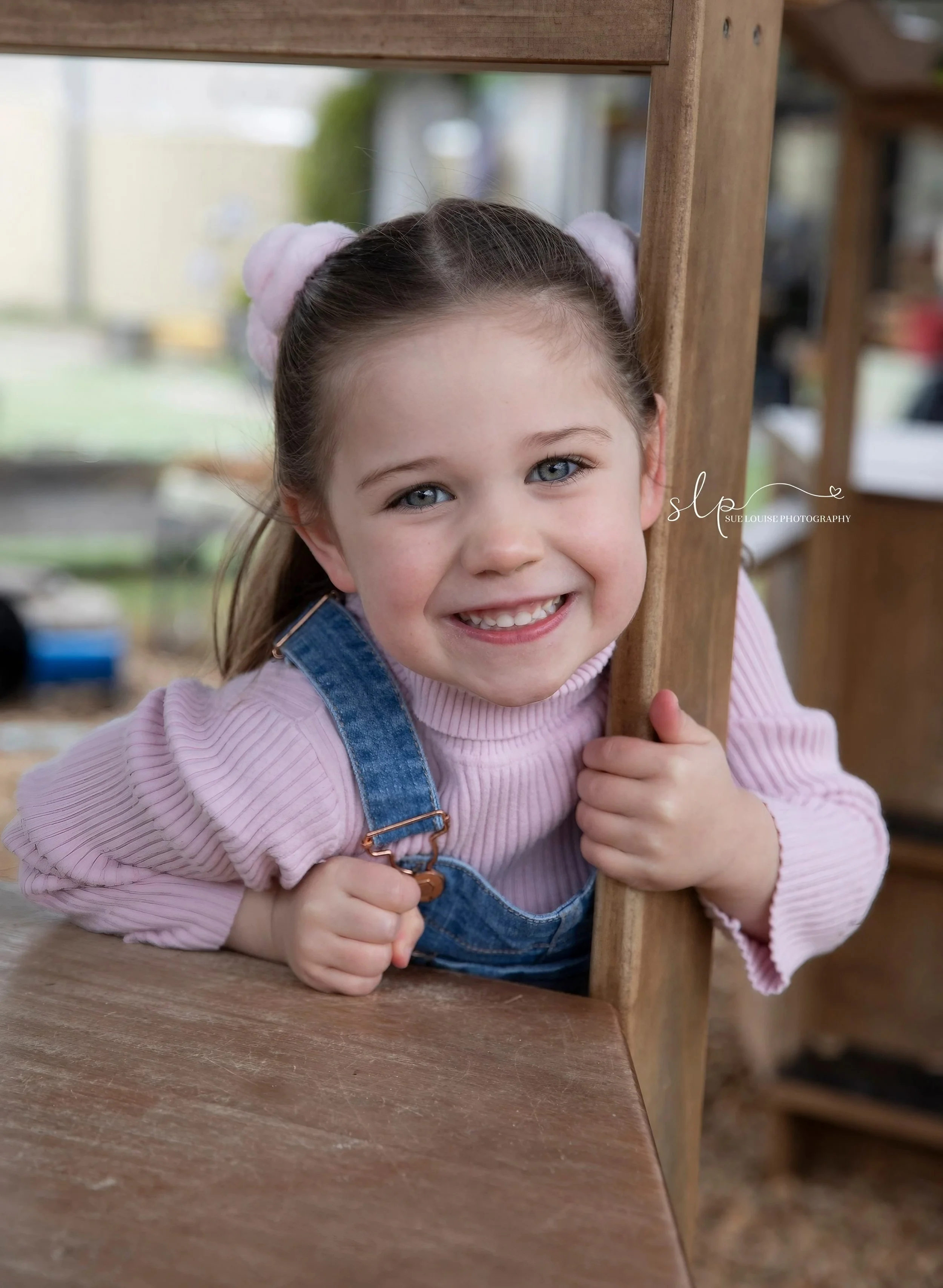A young girl with blue eyes and light brown hair styled in buns with pink hair ties, smiling and peeking through a wooden chair back while holding onto it, wearing a pink sweater and denim overalls, outdoors with a blurred background.