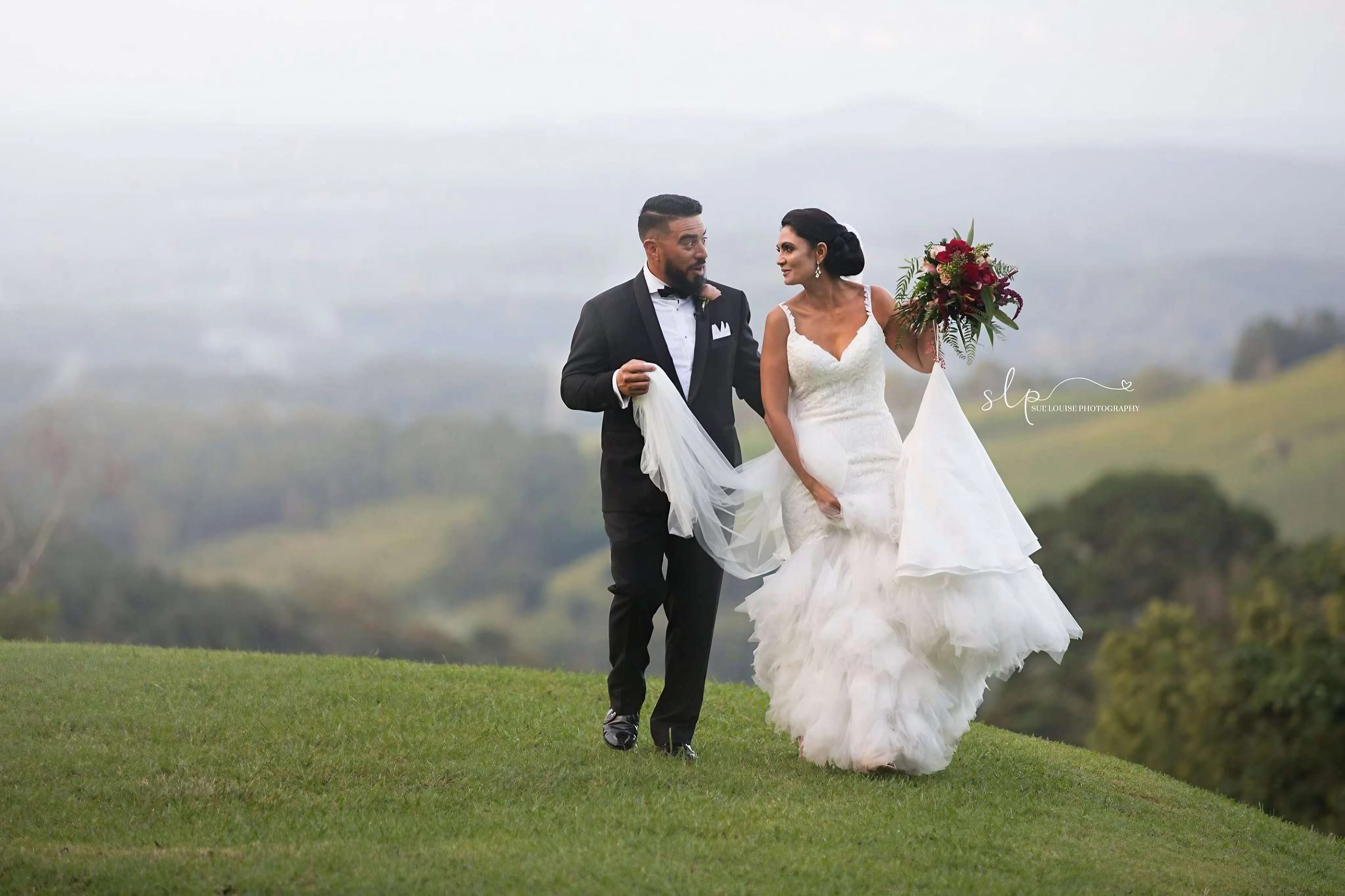 A bride and groom walking on a grassy hilltop, with the bride holding a bouquet of red and pink flowers and wearing a white wedding gown. The groom is dressed in a black tuxedo with a bow tie. The background is foggy with rolling hills and trees.