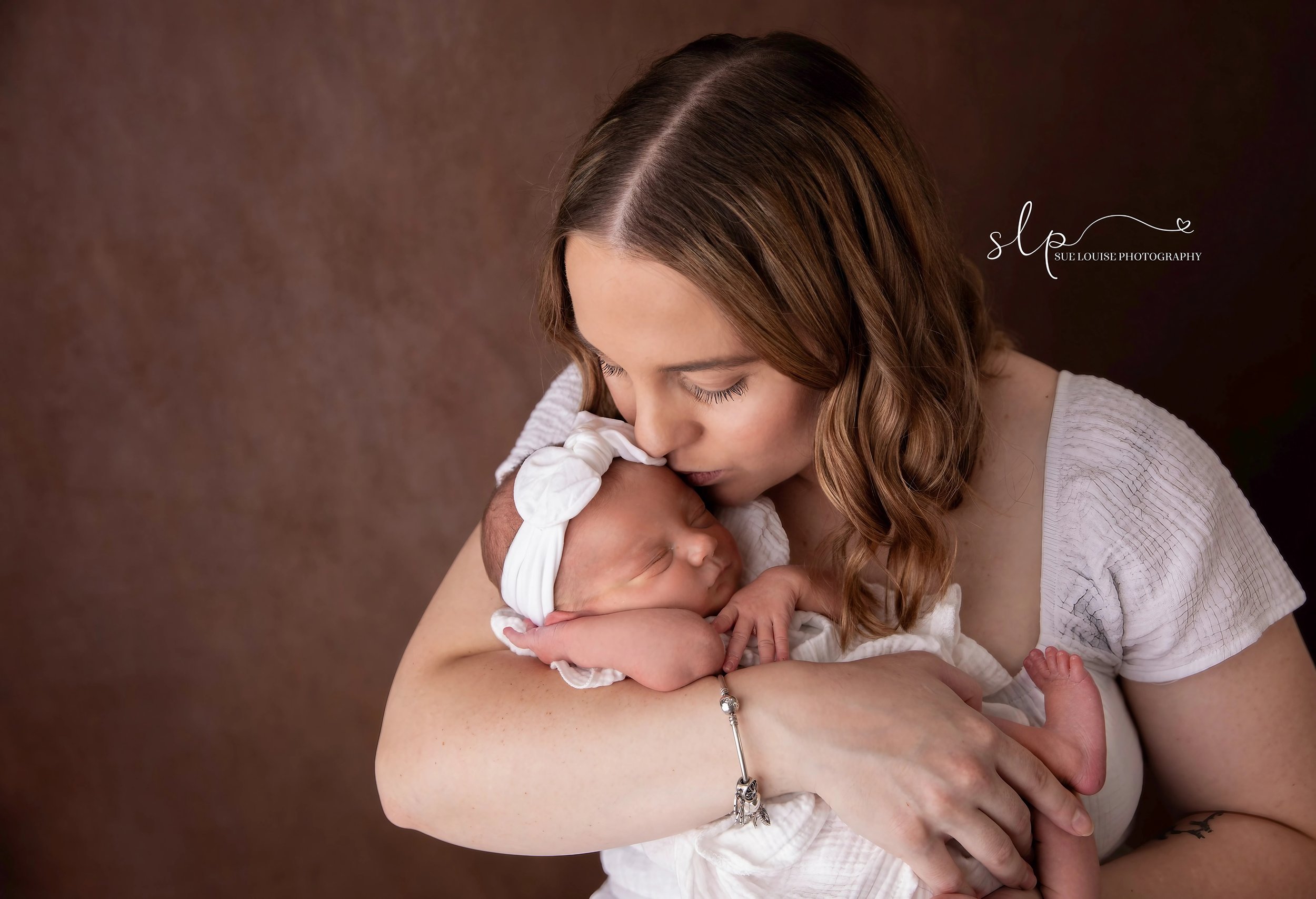 A woman with long brown hair holding a newborn baby with a white headband and outfit, kissing the baby's forehead against her own, against a brown background.