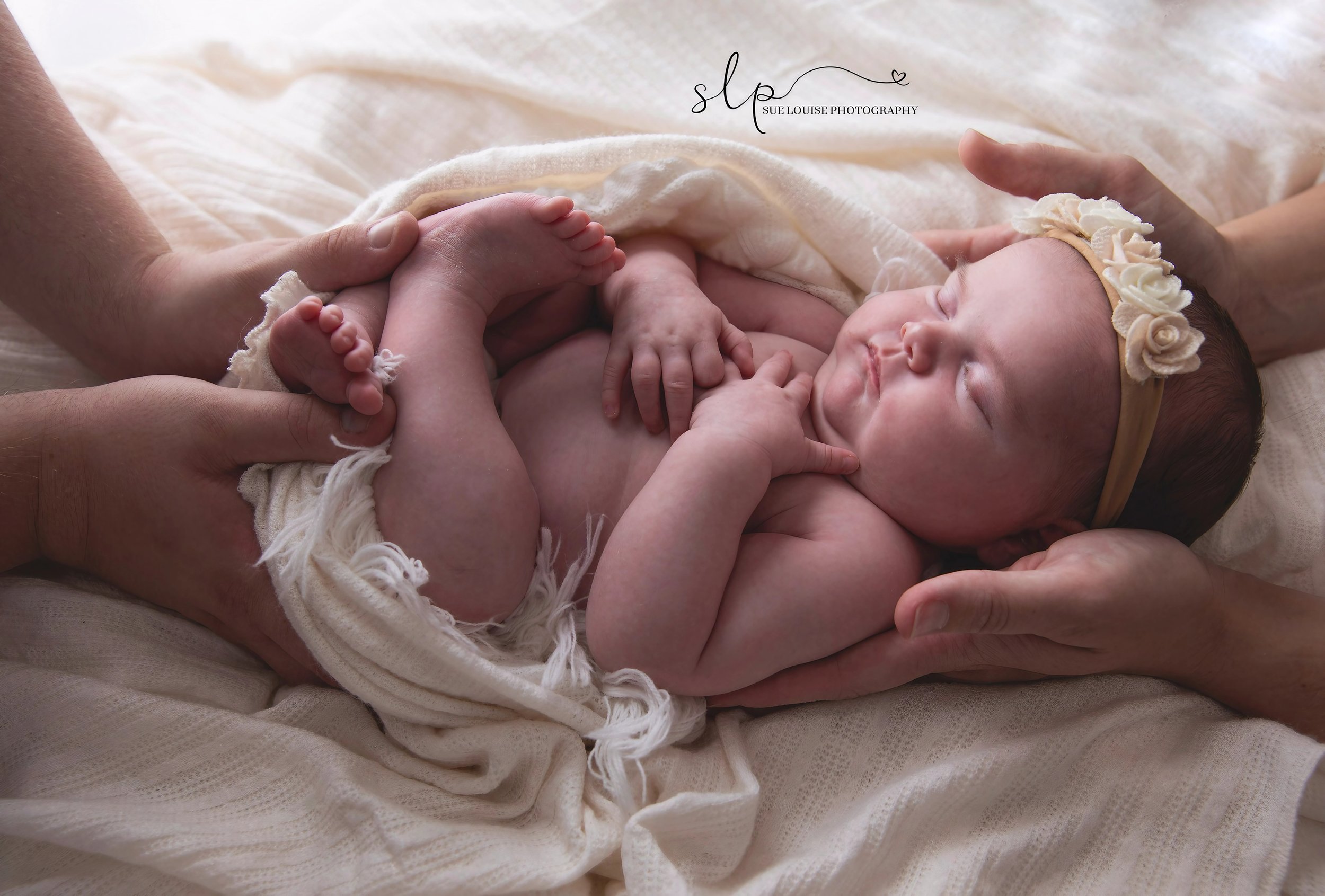 A newborn baby lying on a soft cream blanket, wearing a cream floral headband, with her eyes closed and her arms and legs gently curled. Her mother supports her with both hands.