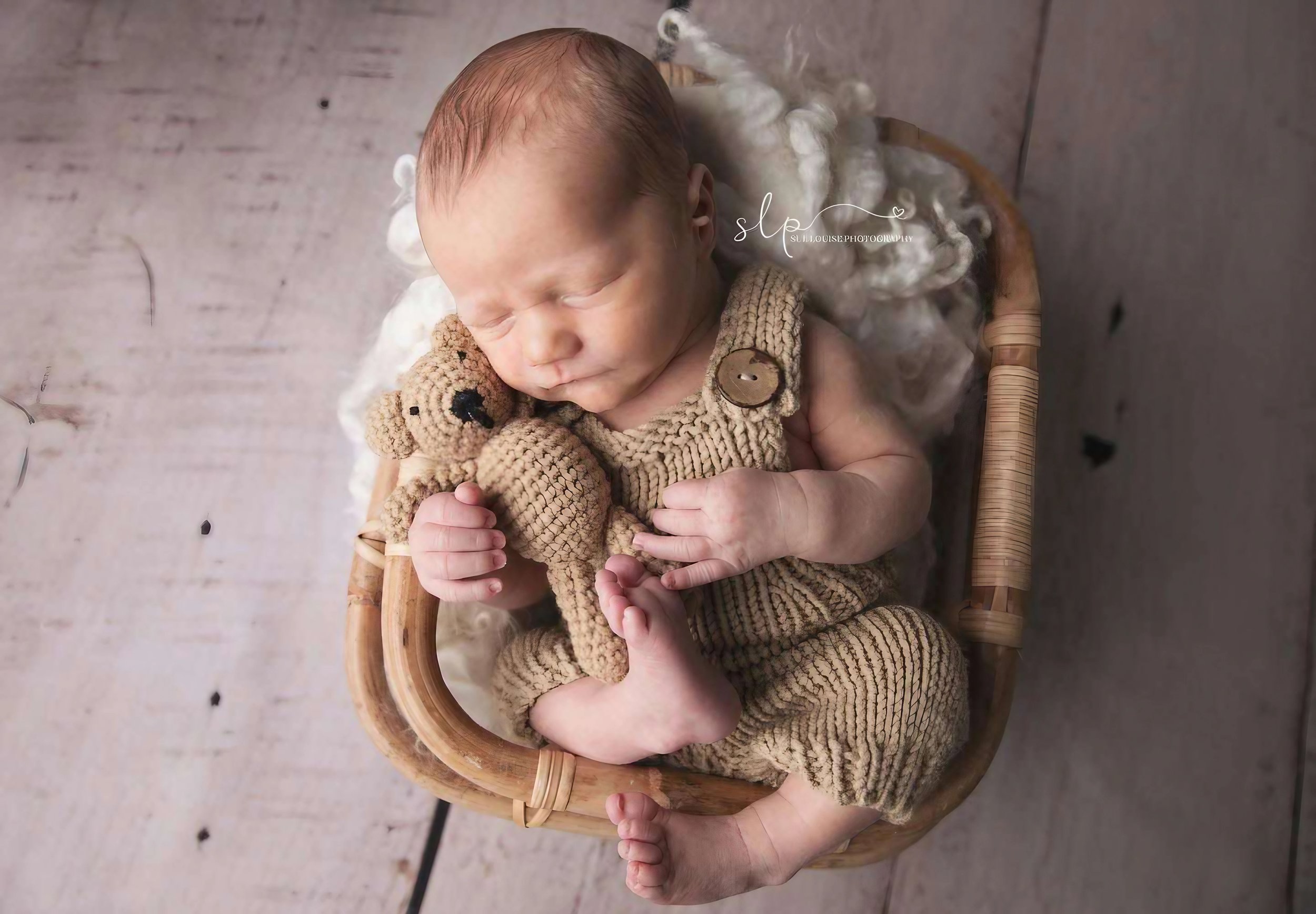 Newborn baby peacefully sleeping in a wicker basket, cuddling a small crocheted teddy bear, on a soft fluffy blanket with a wood floor background.