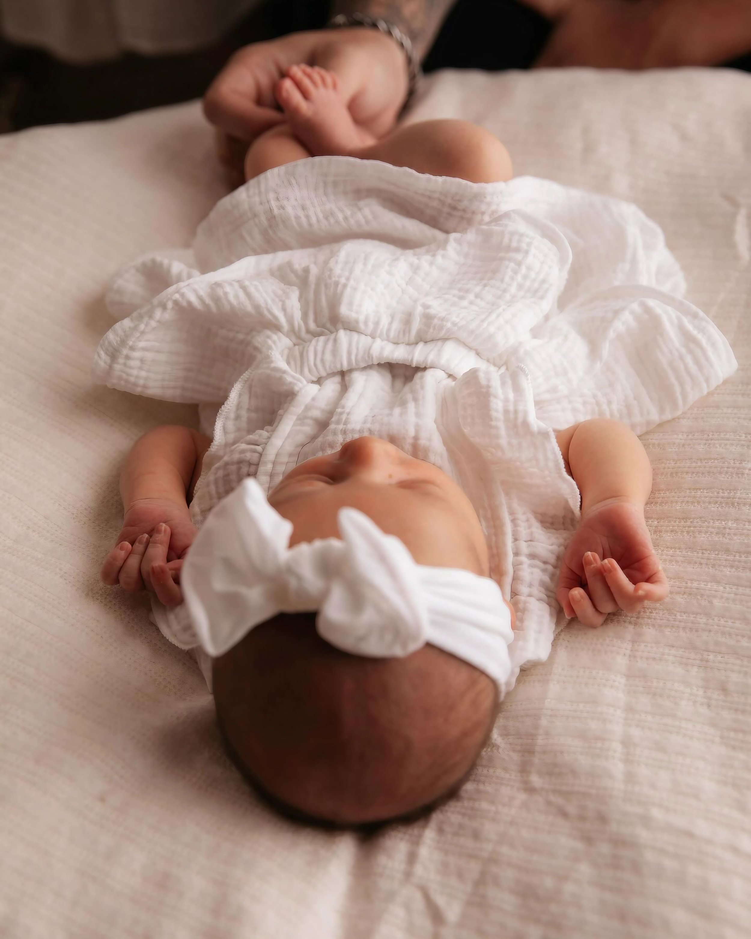A sleeping baby lying on a soft white surface, wearing a white headband and a white textured outfit, with a caregiver's hand gently holding one foot.