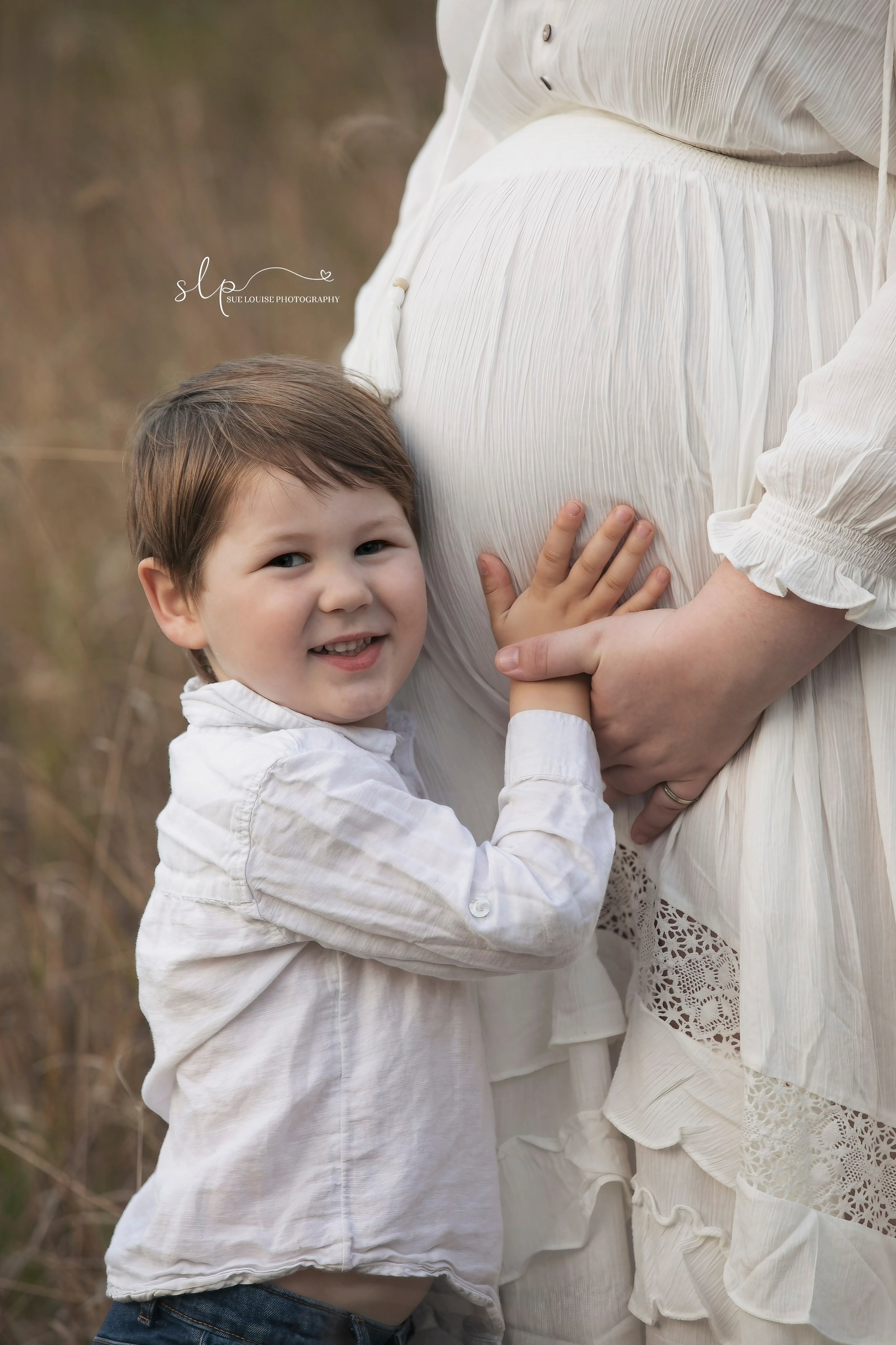 A young boy with brown hair and a white shirt hugging a pregnant woman's belly from the side, outdoors in a natural setting.
