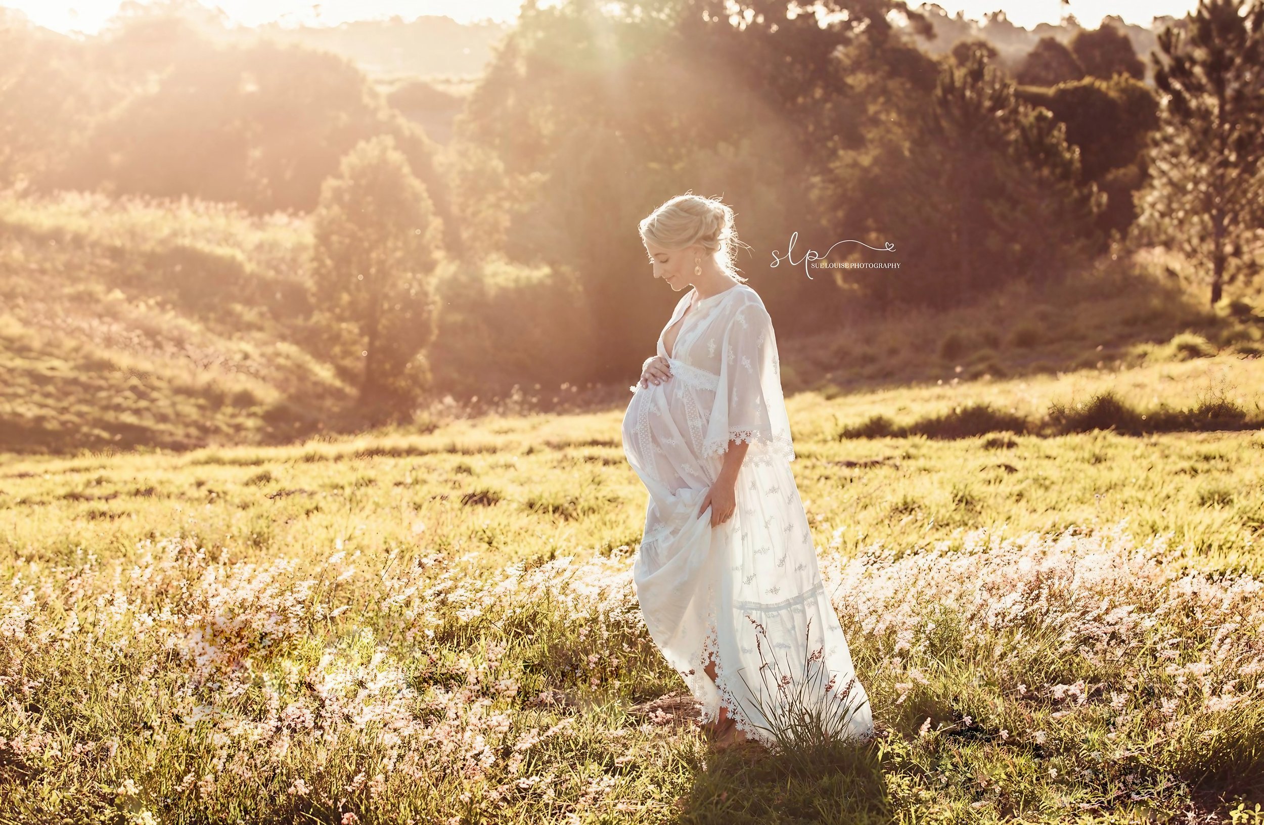 Pregnant woman in a white dress standing in a field of flowers during sunset, with a backdrop of trees and rolling hills.