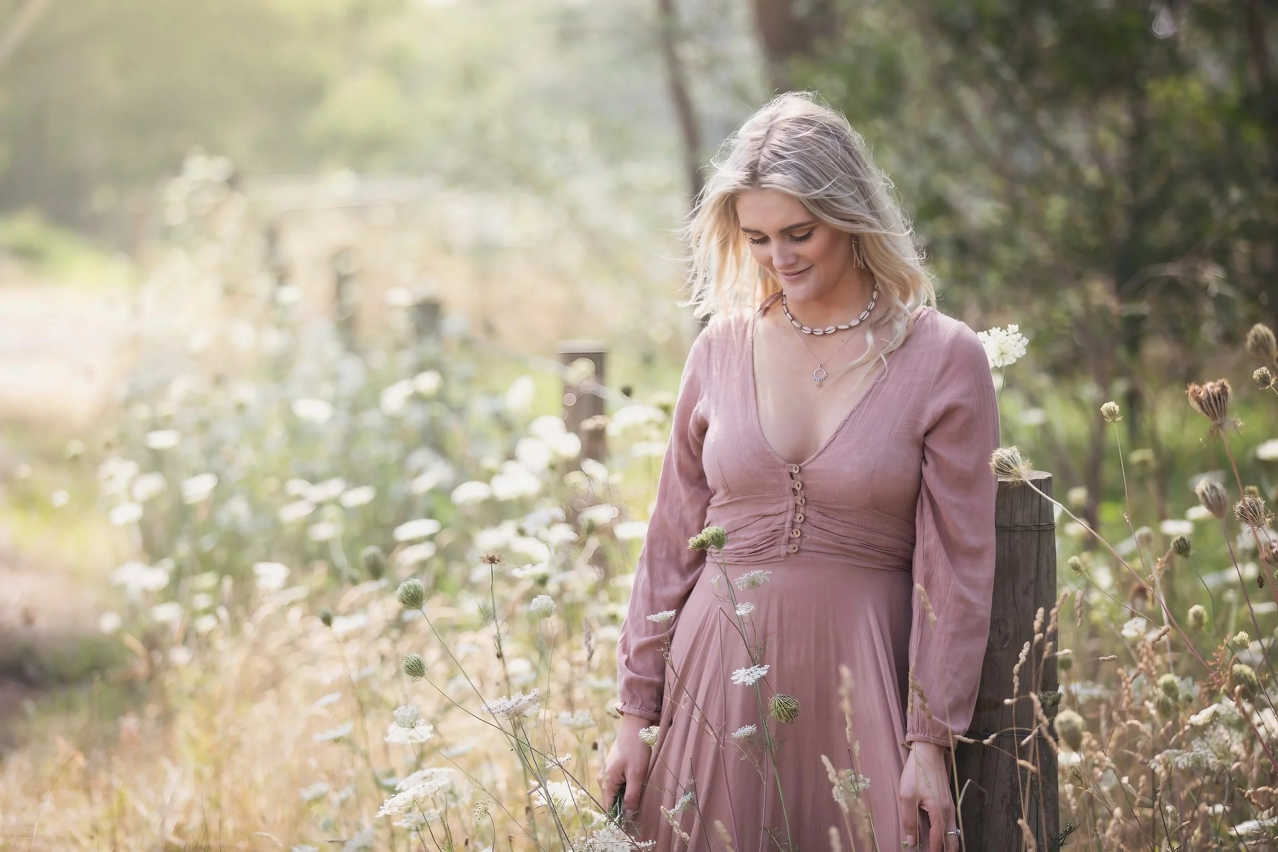 A woman in a long pink dress standing in a field of wildflowers with a wooden fence post nearby, looking downward with a gentle smile.