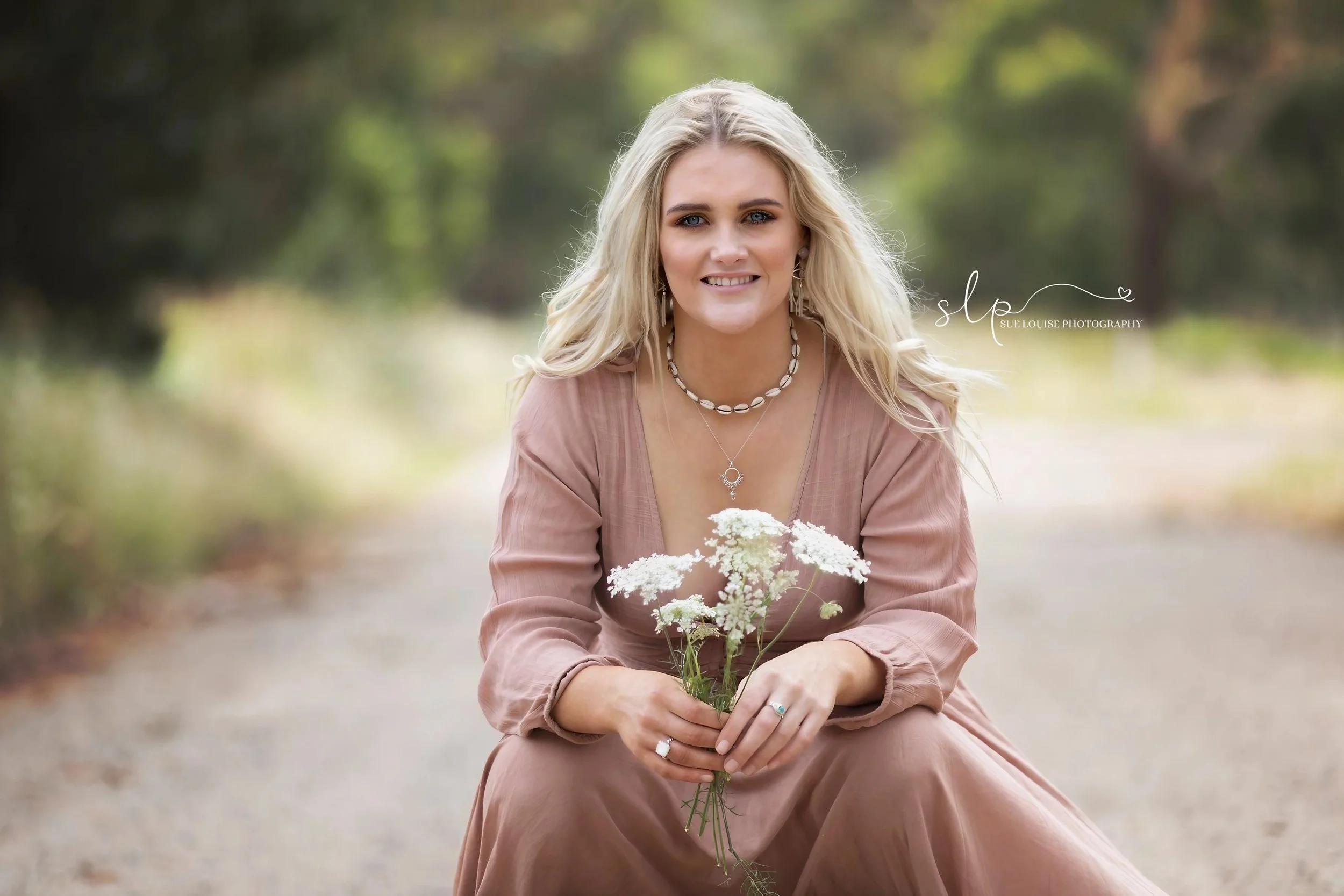 A young woman with wavy blonde hair sitting outdoors on a dirt path, holding a bouquet of white flowers, wearing a brown dress, jewelry, and smiling at the camera with a blurred natural background.