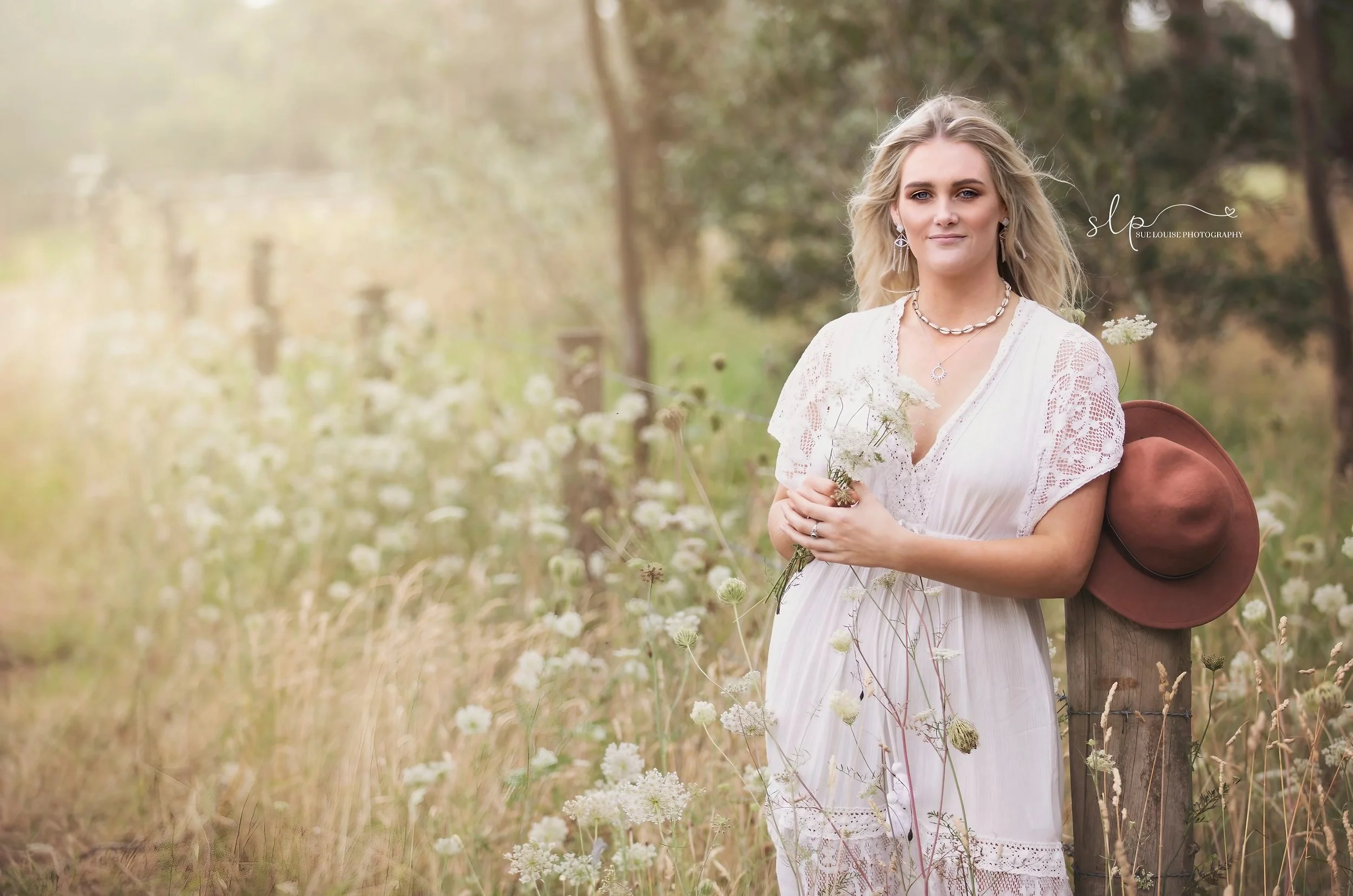 A woman in a white dress holding flowers in a field of tall grass and wildflowers, with a wide-brimmed brown hat resting on a wooden post beside her.