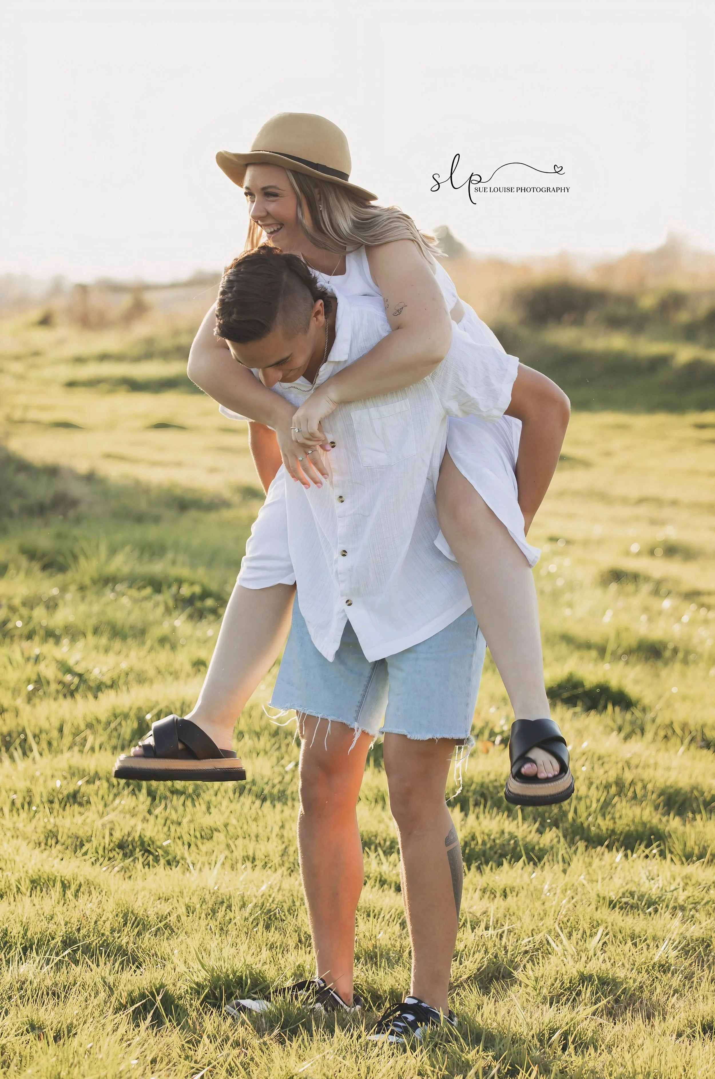 A smiling woman wearing a beige hat and white dress is giving a piggyback ride to a man in a white shirt and denim shorts in a grassy outdoor field during sunset.