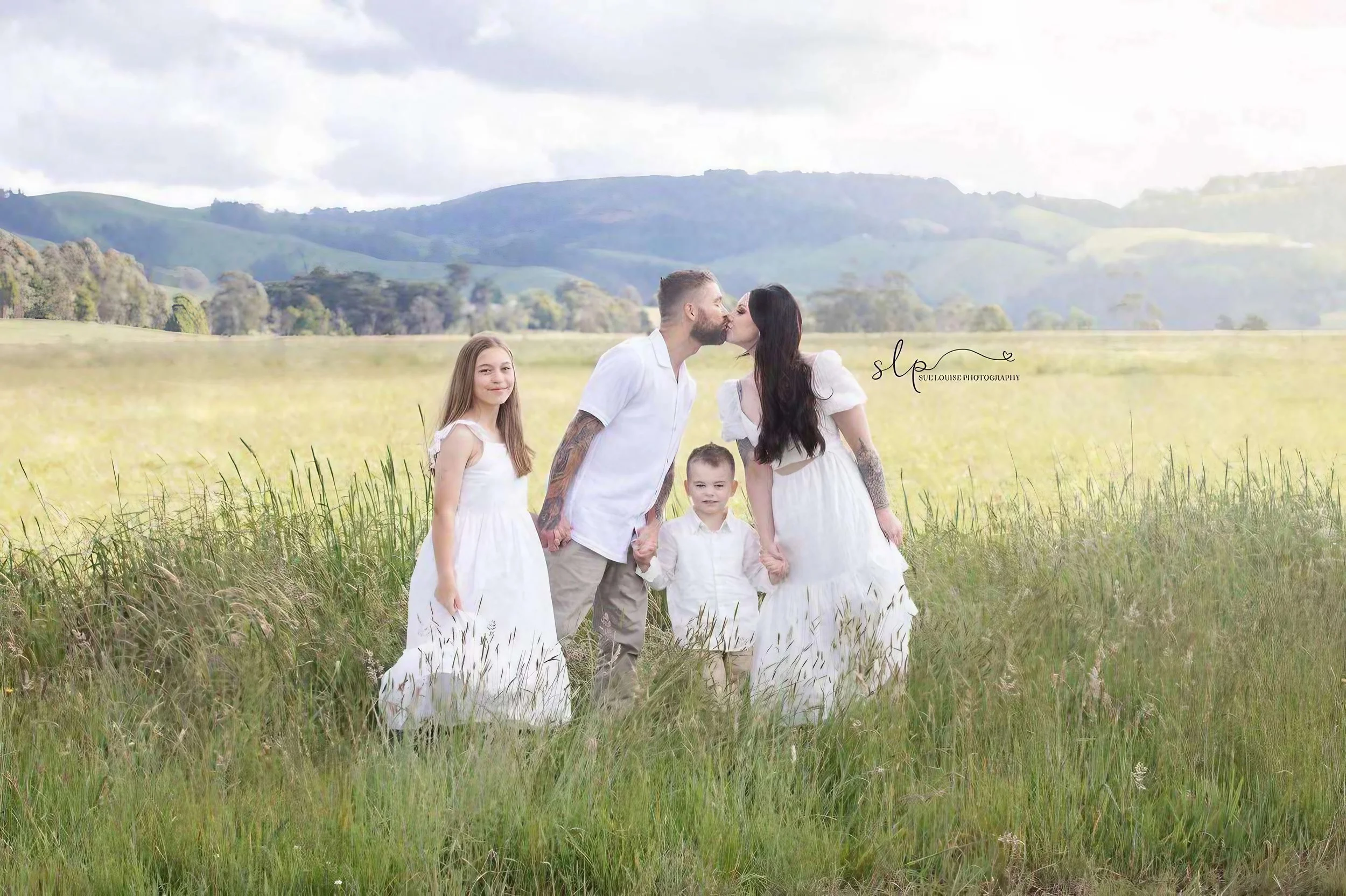 A family of five standing in a grassy field with rolling hills in the background; the two adults kiss while two children stand nearby, holding hands.