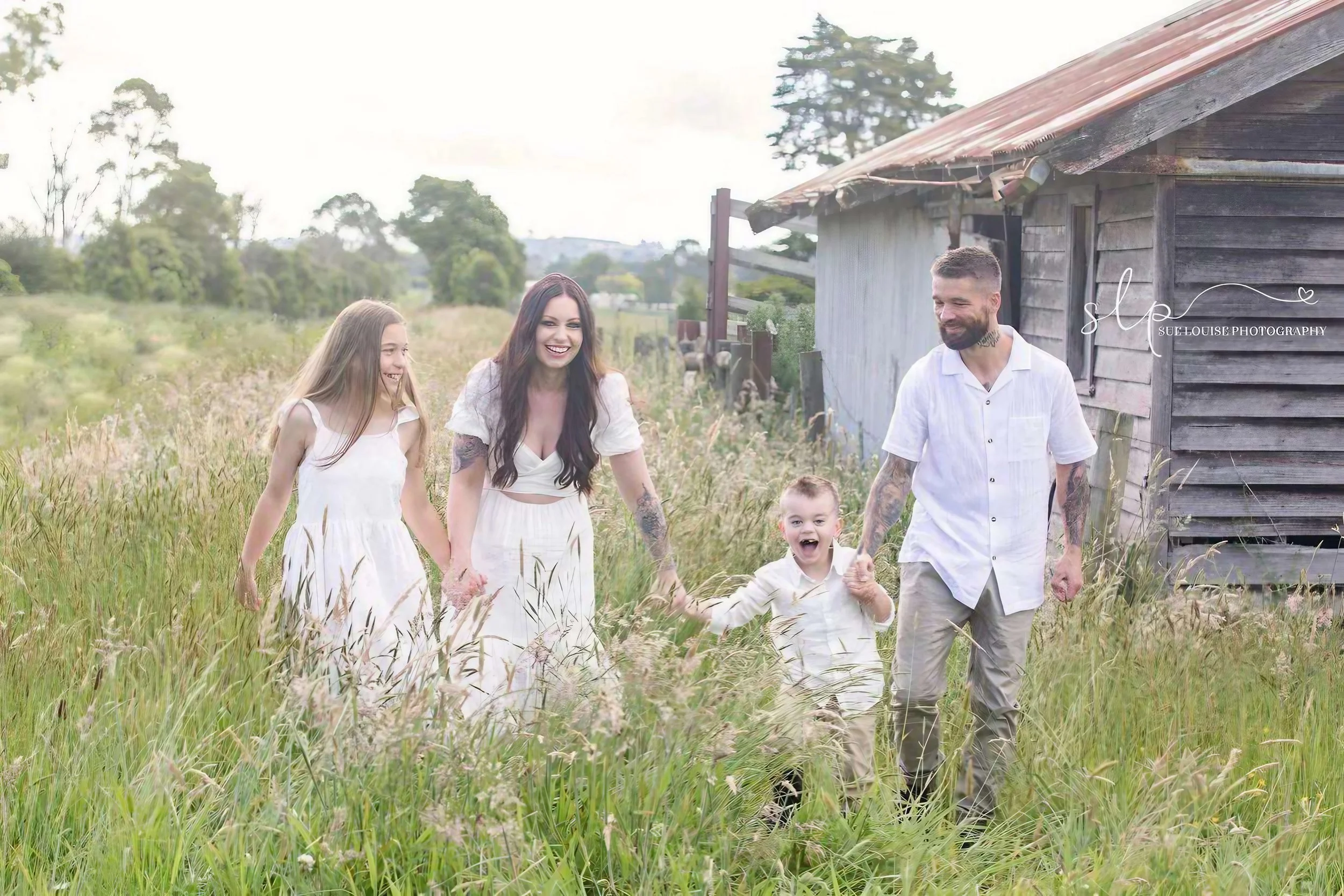 A family of five walking through tall grass near a rustic wooden building, all smiling and holding hands.