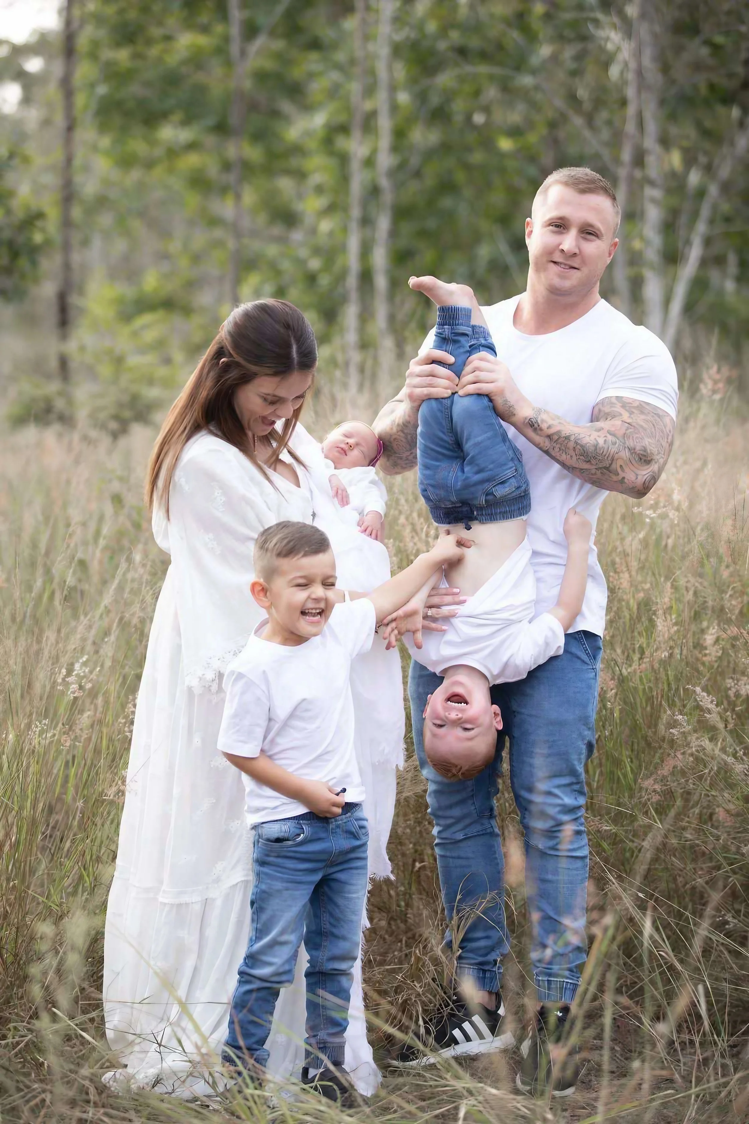 Family of five standing in a field, with a woman holding a newborn baby, a man lifting a boy upside-down, and another boy playing, all smiling and enjoying outdoor time.
