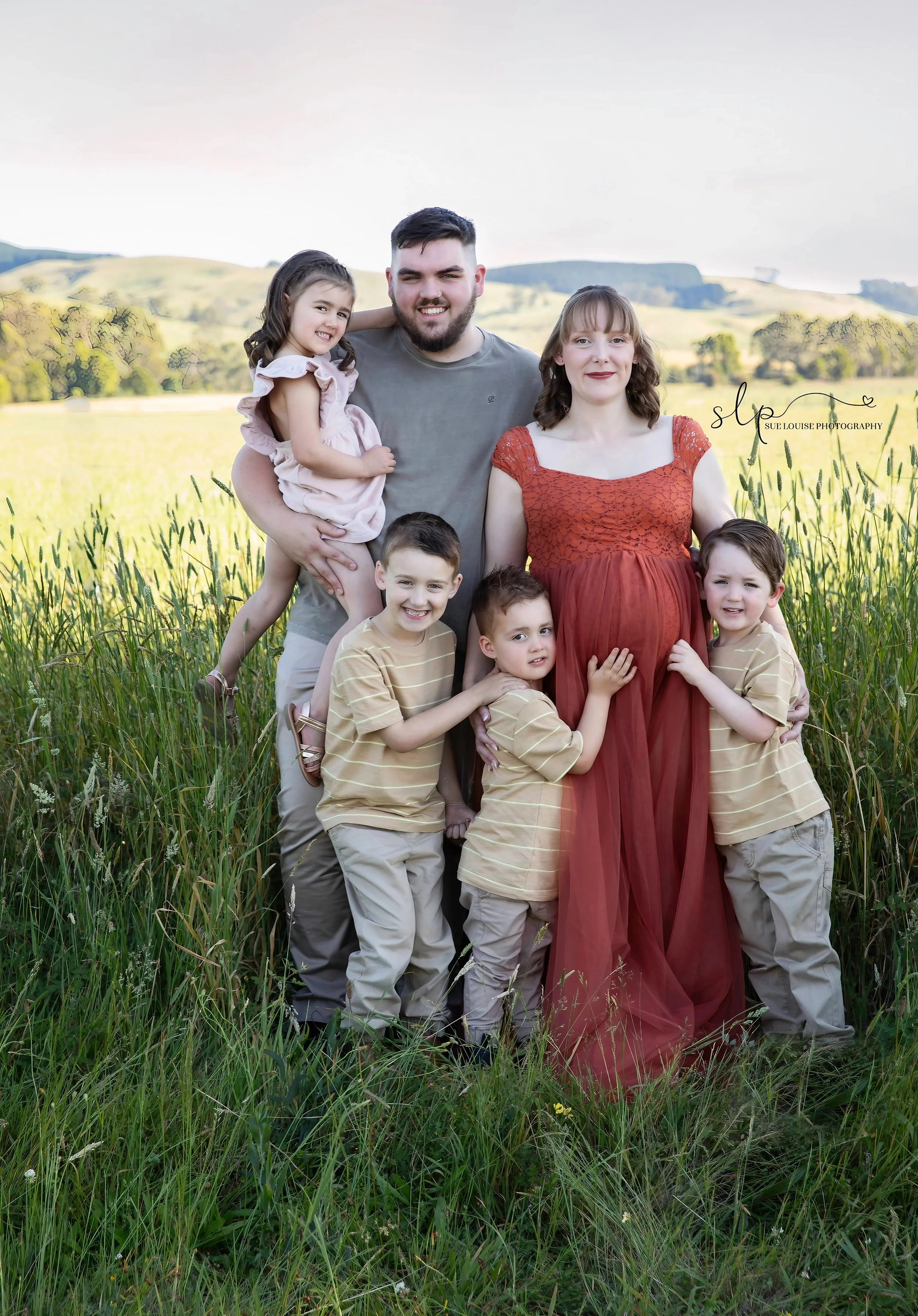 Family portrait in a field with green grass and rolling hills in the background, including two adults and four children.