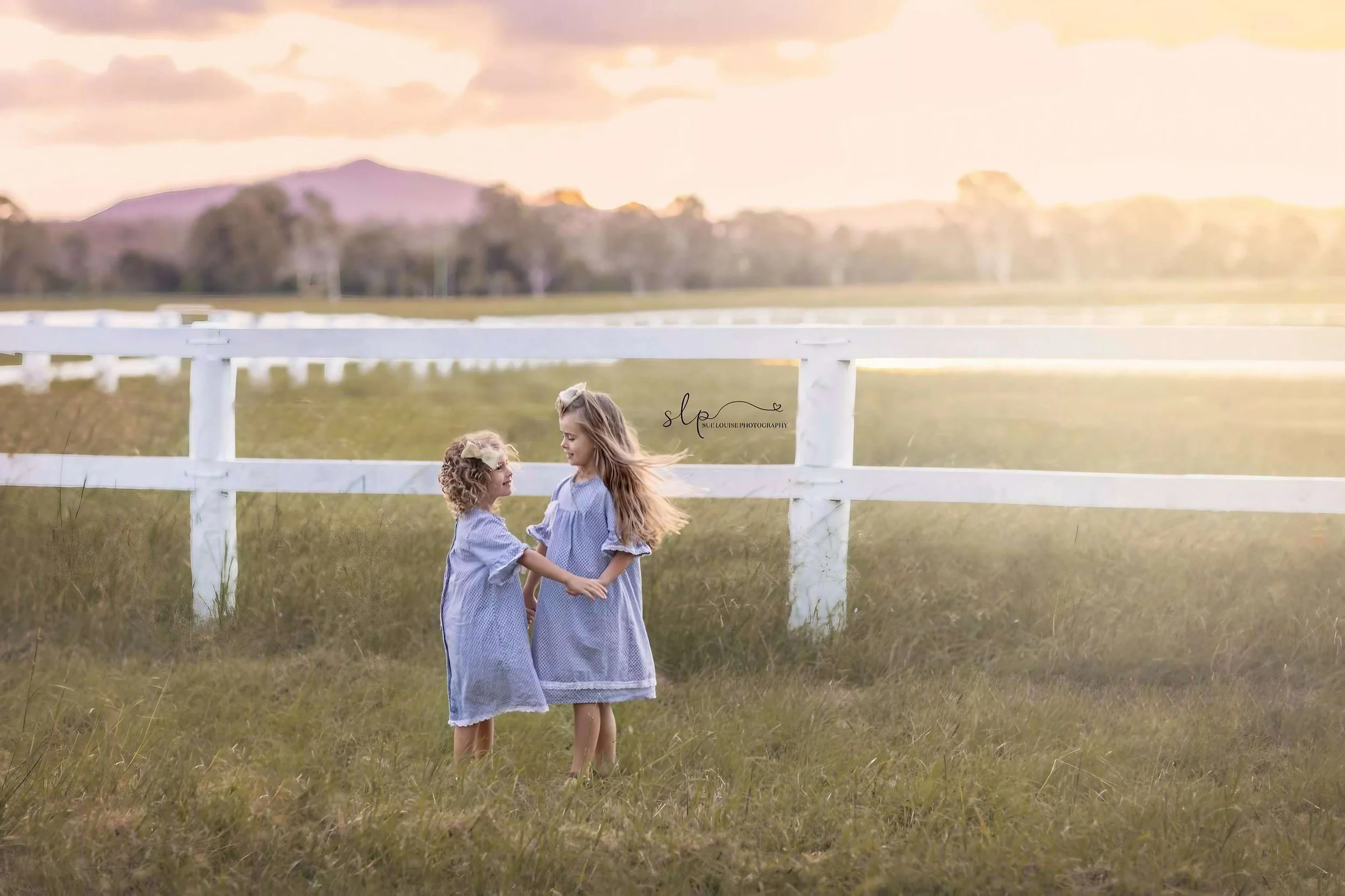 Two young girls wearing matching blue gingham dresses holding hands and looking at each other in a grassy field at sunset, with a white fence behind them and a mountain in the distance.
