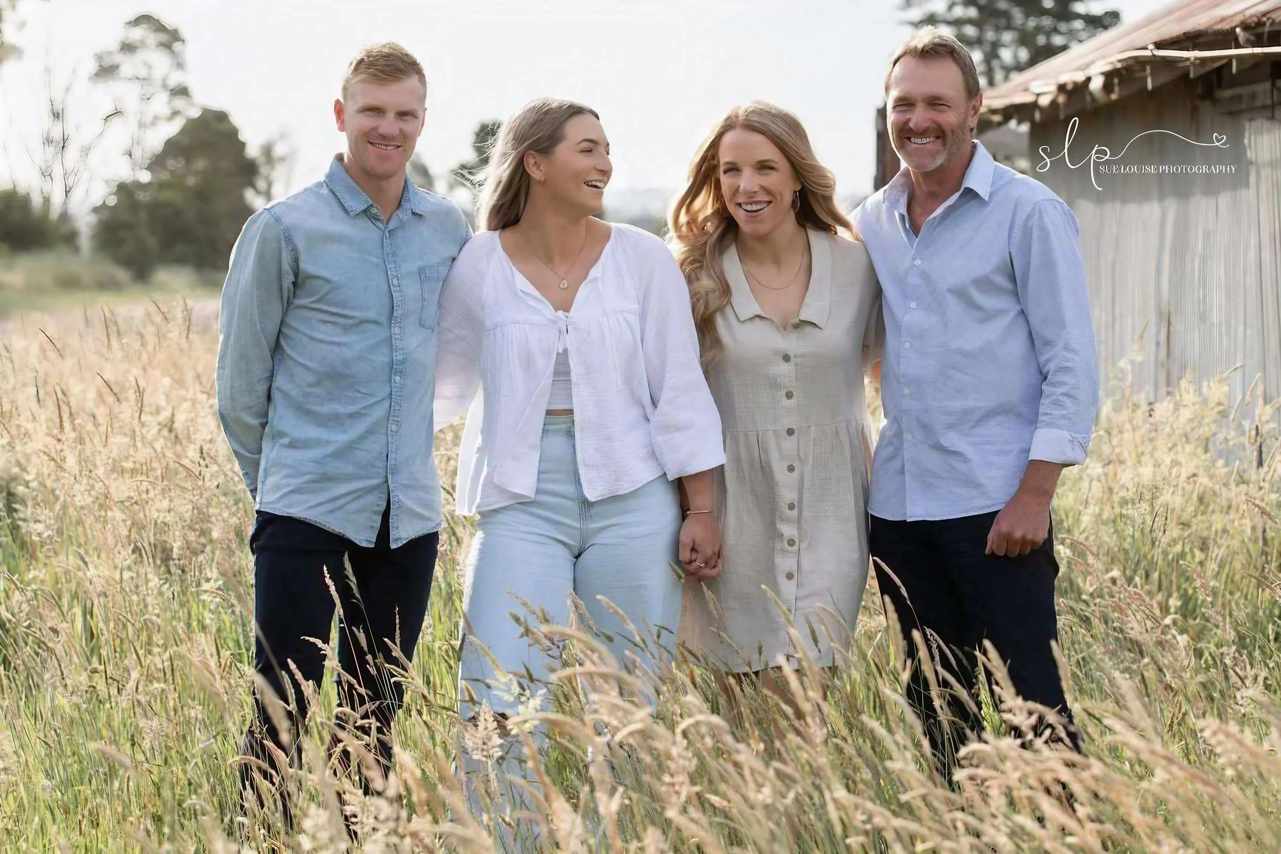 Four people holding hands and smiling in a grassy field with a rustic barn in the background, during daytime.