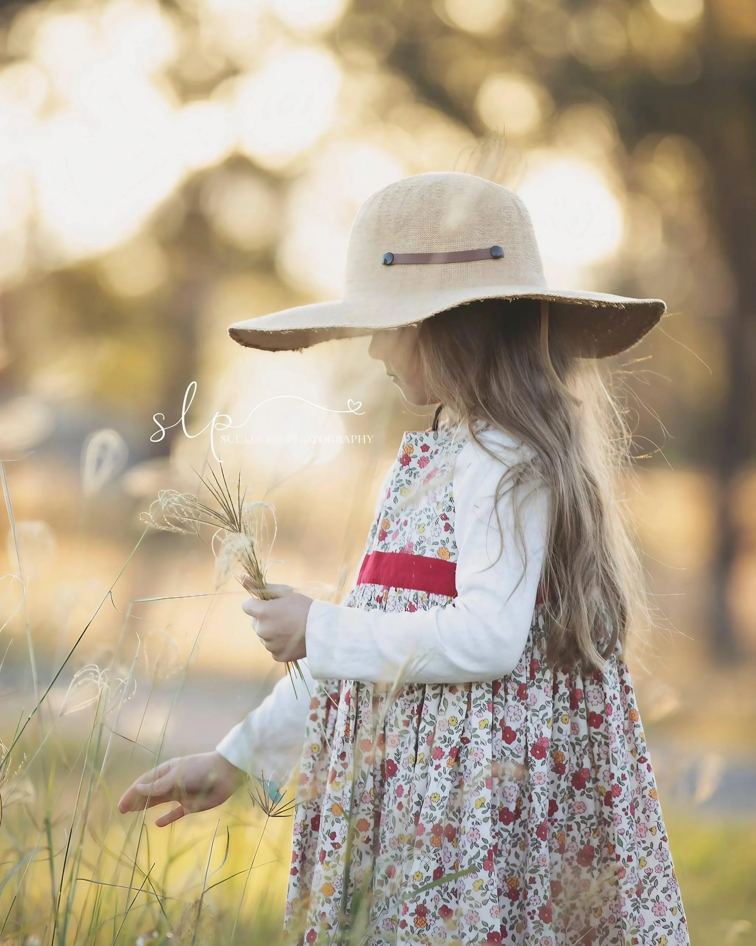 A young girl in a floral dress and cream-colored long-sleeve shirt holding a small bunch of wildflowers, wearing a large floppy sunhat, outdoors in a field during golden hour.