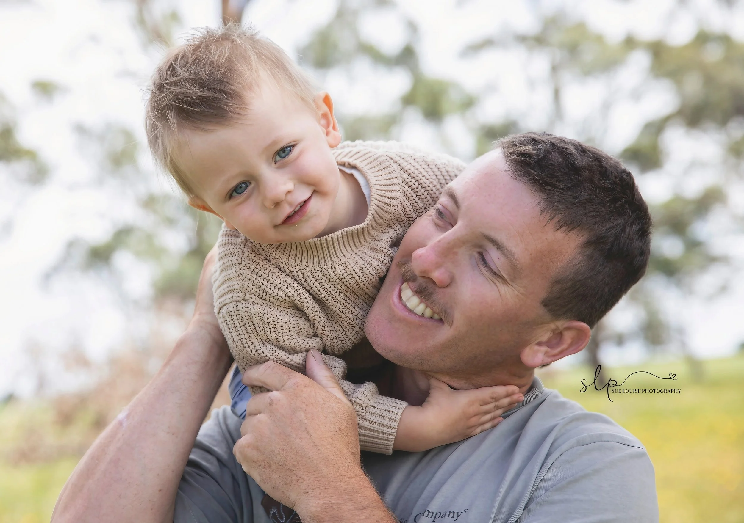 A man playfully holding a young boy on his shoulders outdoors. The boy is smiling and wearing a beige sweater, while the man is smiling and looking at the boy. Trees and a bright sky are blurred in the background.